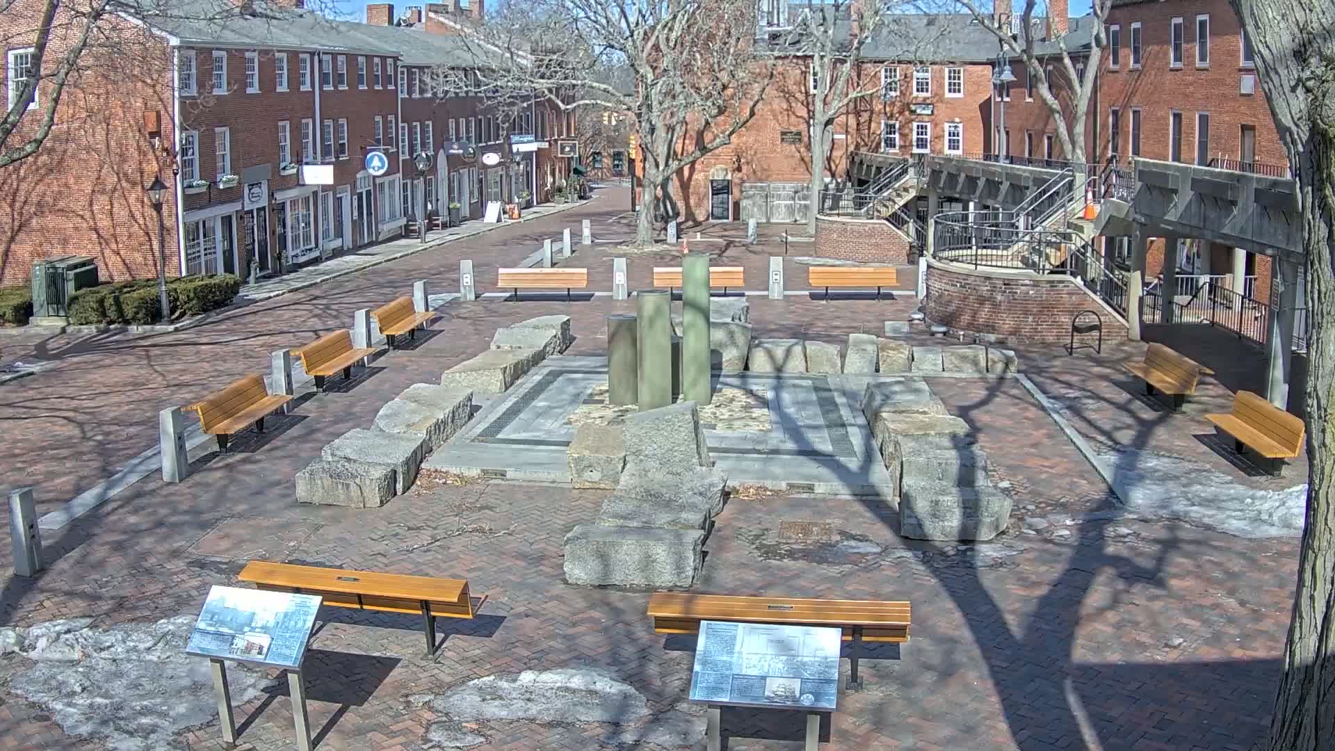 A brick plaza with several benches, a central stone structure, and a modern fountain, surrounded by brick buildings under a sunny sky.