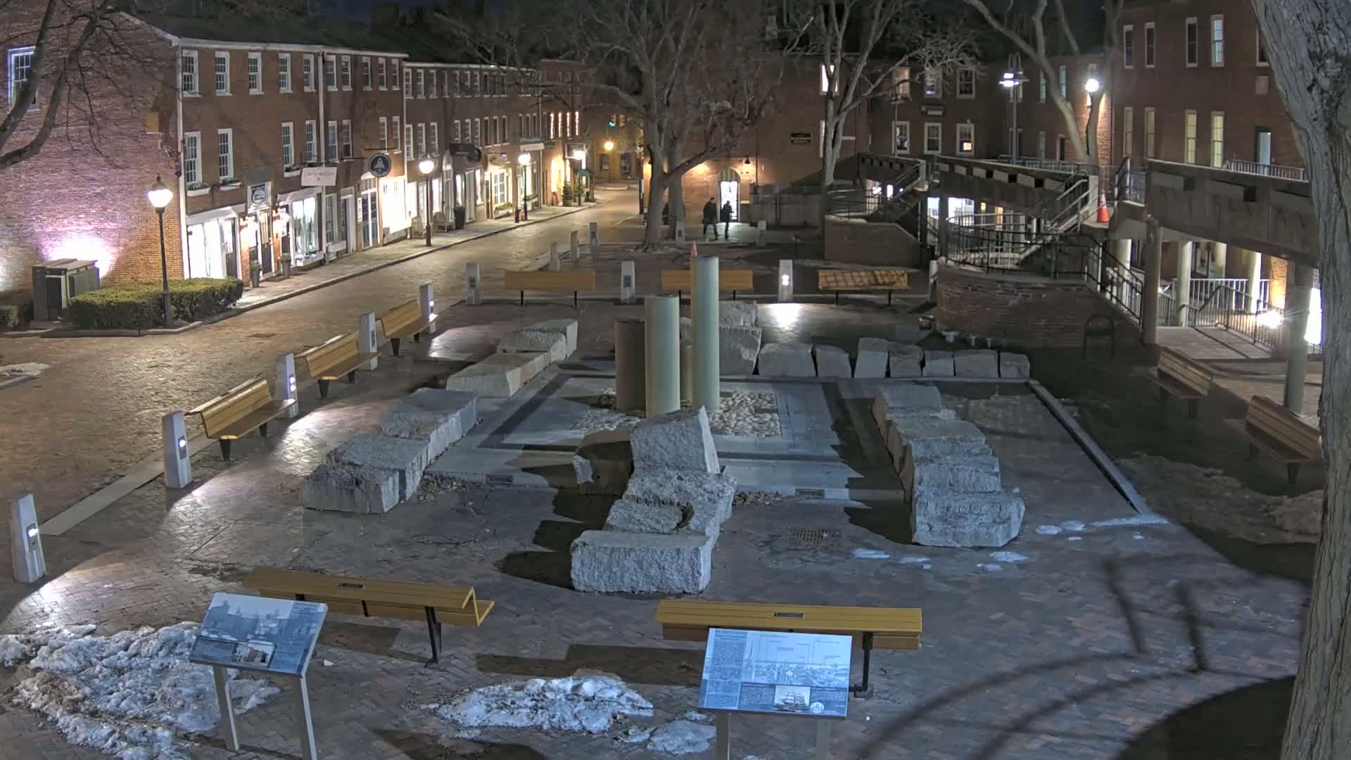 At night, a brick-paved town square features a stone structure, several benches, and trees with bare branches, under dark, clear skies.
