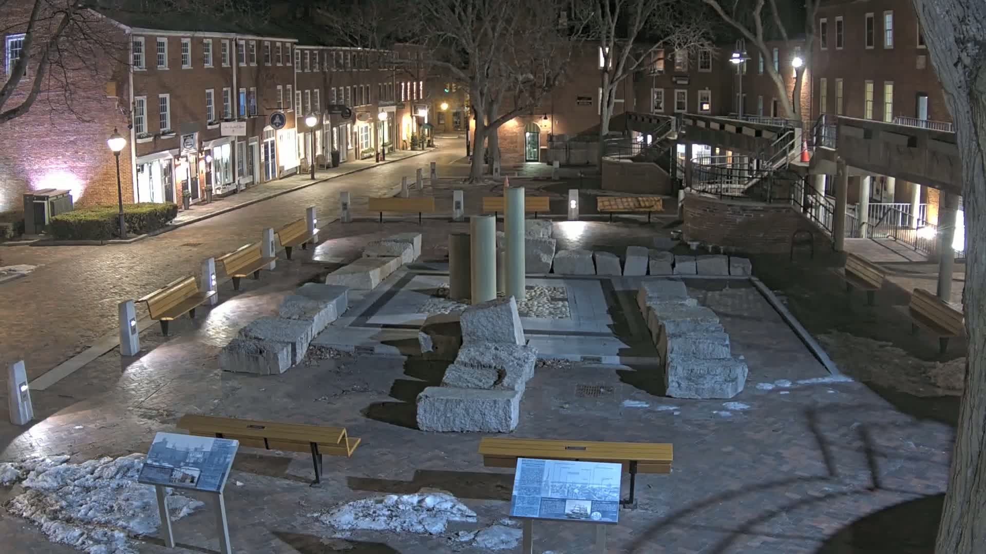 At night, a brick-lined square contains the ruins of a stone structure, surrounded by benches and illuminated by streetlights.