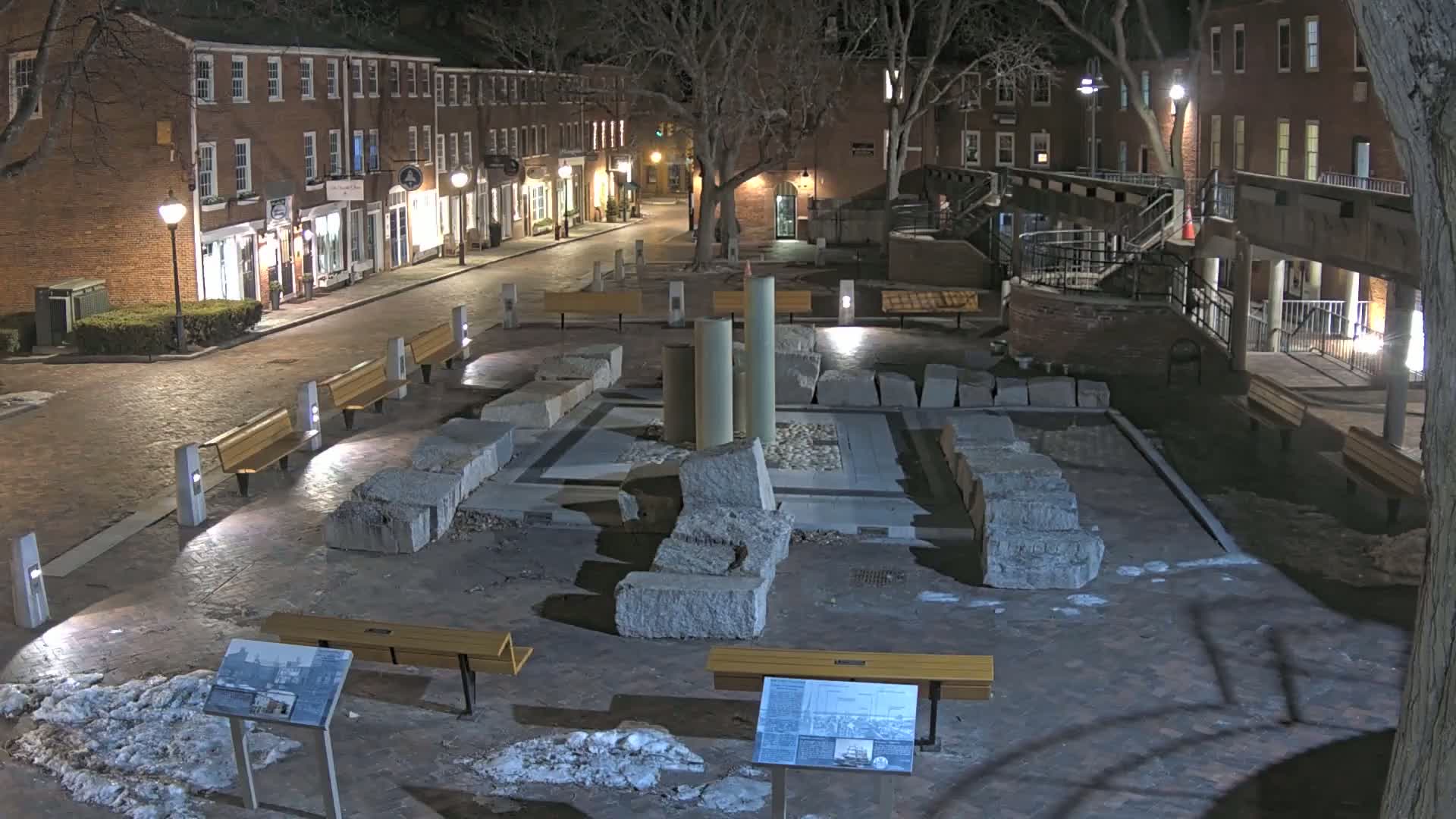 A nighttime scene shows a plaza with remnants of stone columns and walls, surrounded by benches and brick buildings lit by streetlights, with patches of snow on the ground.