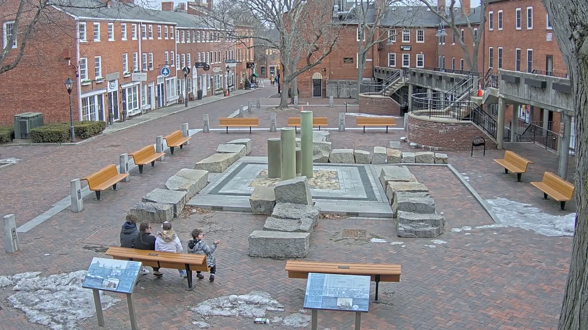 A brick plaza with benches and a stone fountain is surrounded by brick buildings, with patches of snow and ice on the ground.