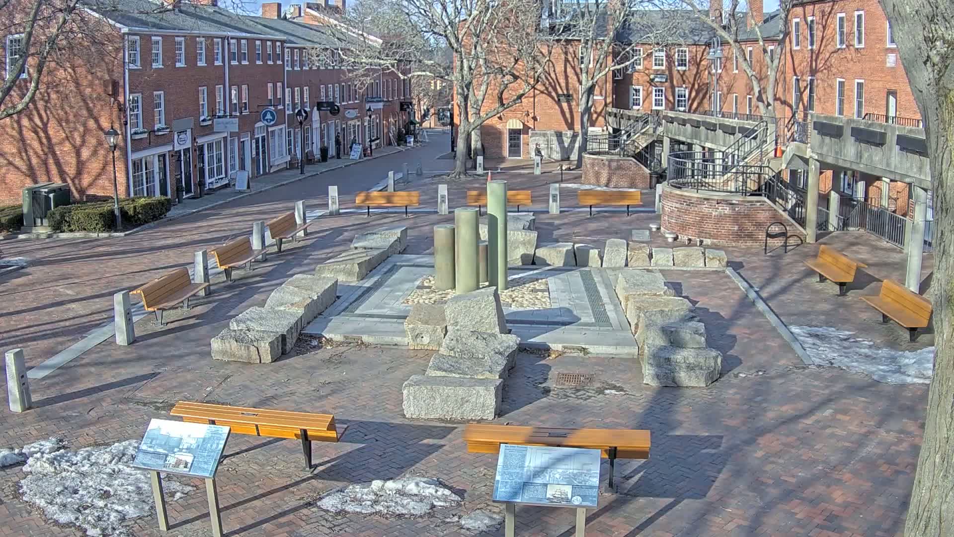 A brick plaza with several benches surrounds a stone fountain-like structure, on a sunny day with patches of snow and ice on the ground.