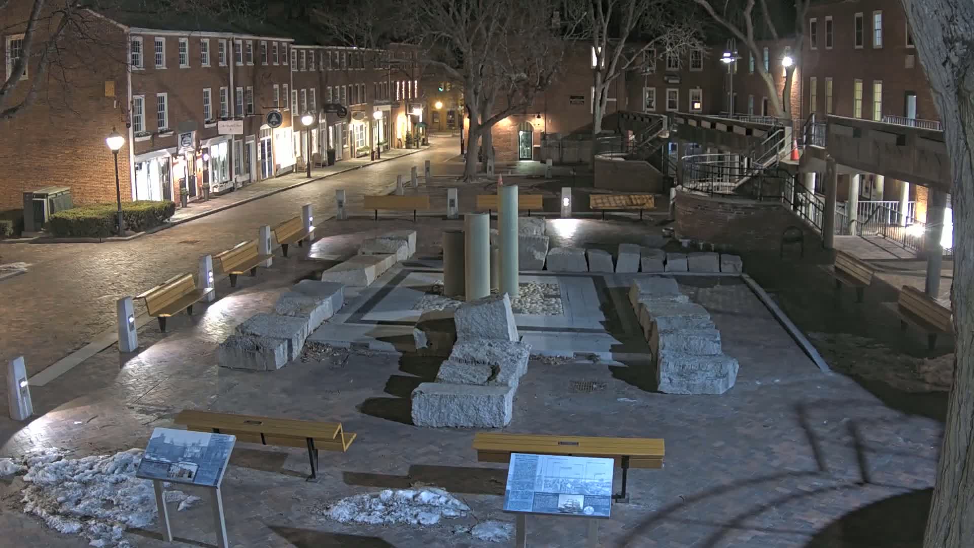 At night, a brick-lined plaza features remnants of a stone structure in the center, surrounded by benches and illuminated by streetlights; small patches of snow are present.