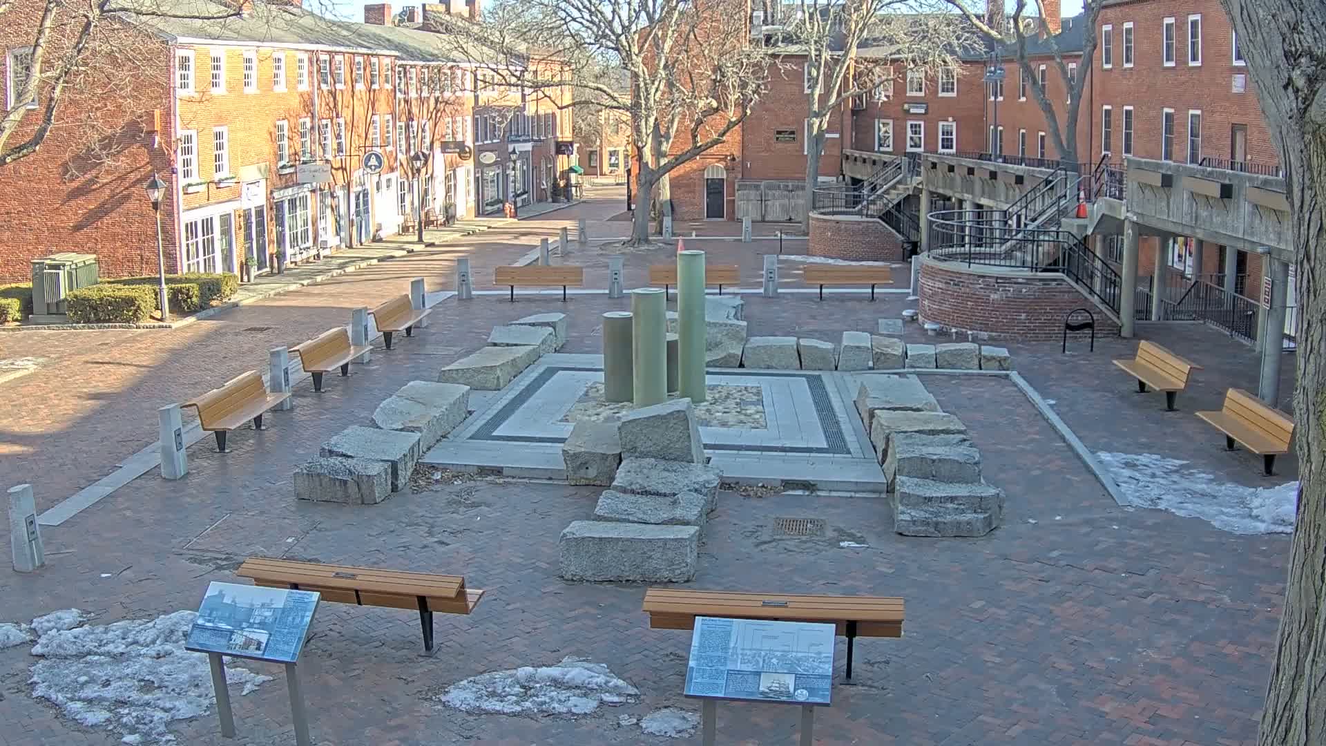 A brick plaza with several benches surrounds a central stone structure and several vertical columns under a sunny winter sky with patches of snow on the ground.