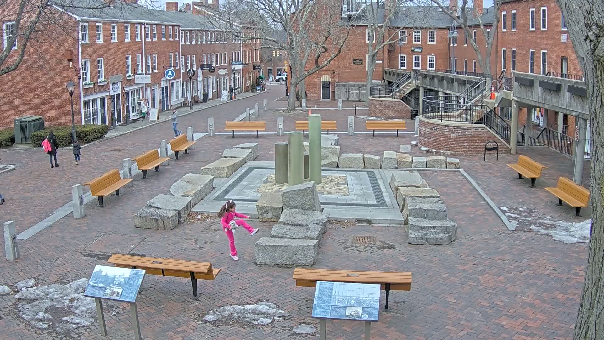 A brick plaza with several benches and a stone fountain is surrounded by brick buildings on a partly cloudy day.