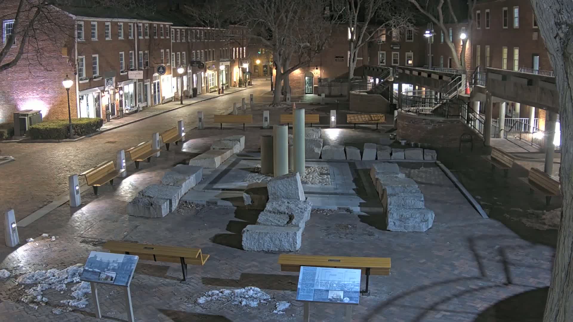 At night, a brick-paved plaza contains a stone ruin display, surrounded by benches and lit by streetlights, with some snow on the ground near the buildings lining the street.