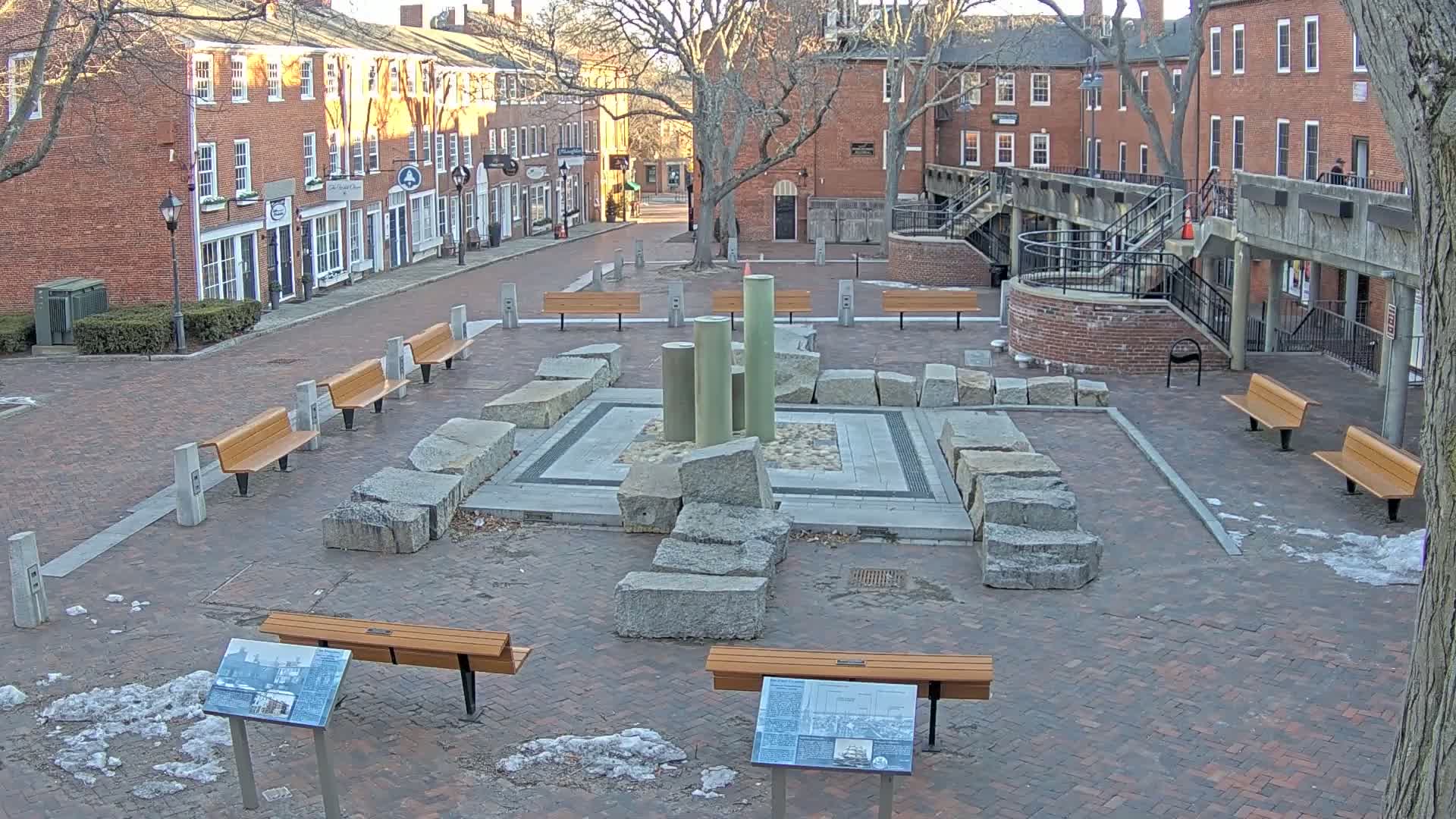 A brick plaza with several benches and a stone fountain is situated between brick buildings on a partly sunny, cold day with patches of snow on the ground.