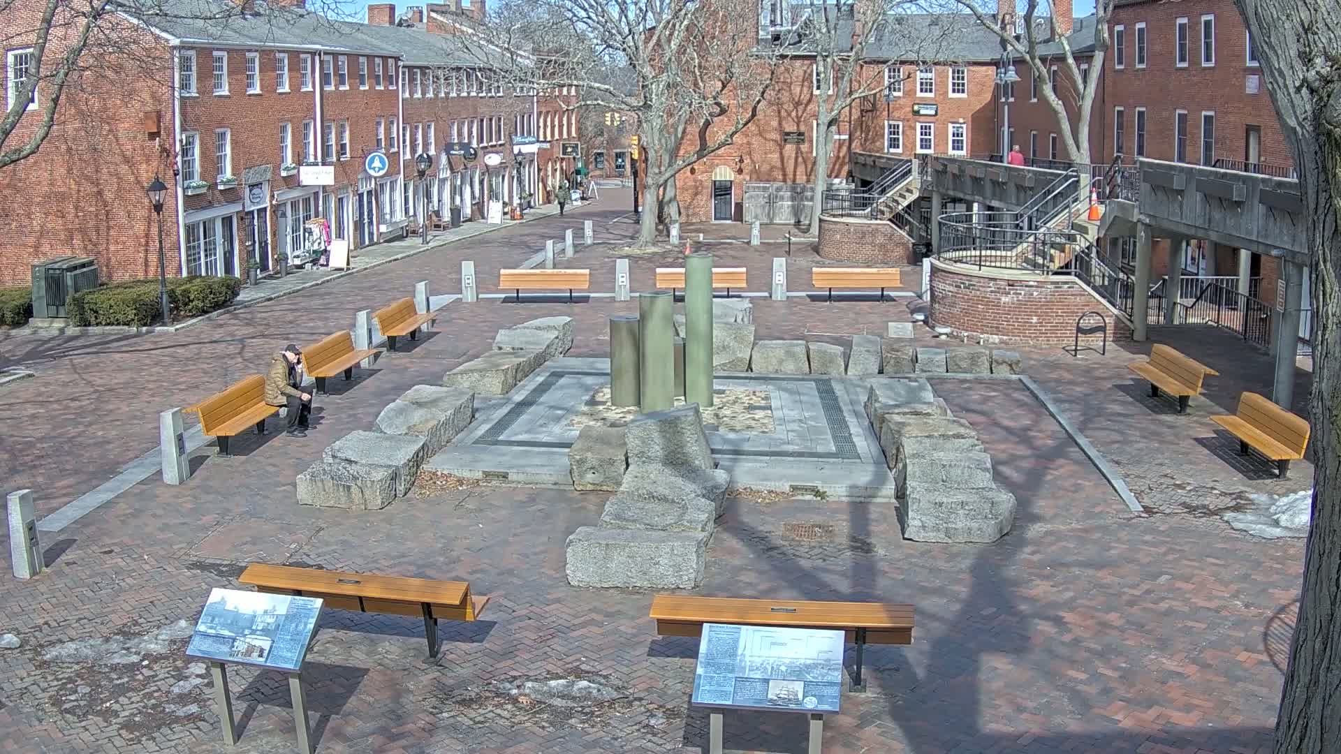 A brick-paved town square with benches, a central stone and metal fountain feature, and surrounding brick buildings on a sunny day.