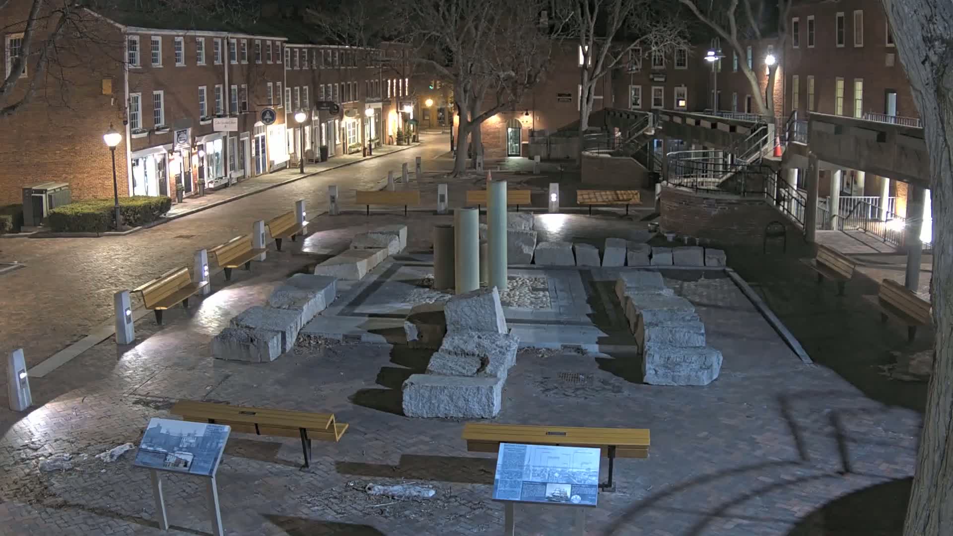 At night, a brick-paved plaza contains the ruins of a stone structure, several benches, and streetlights illuminating the area.