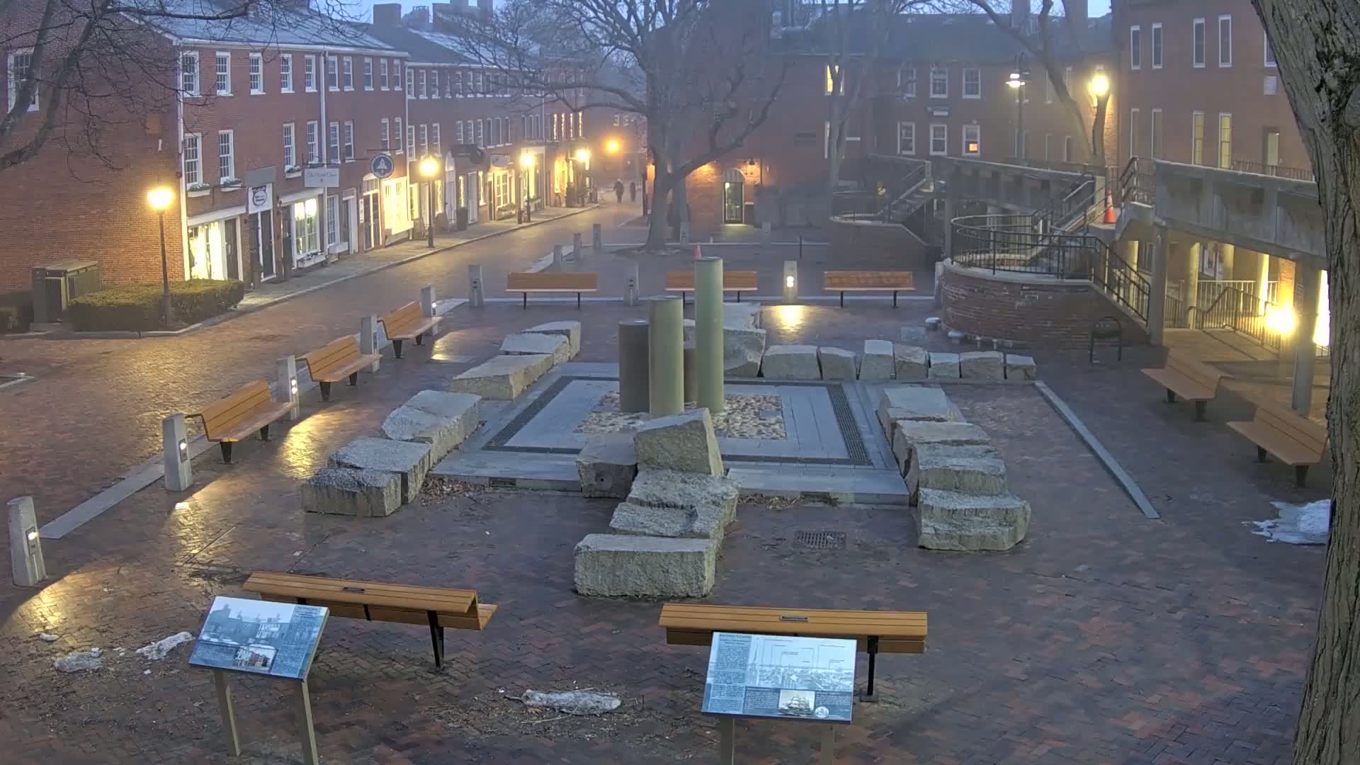 A brick plaza at dusk, featuring several benches, a stone fountain, and surrounding buildings with lights on.