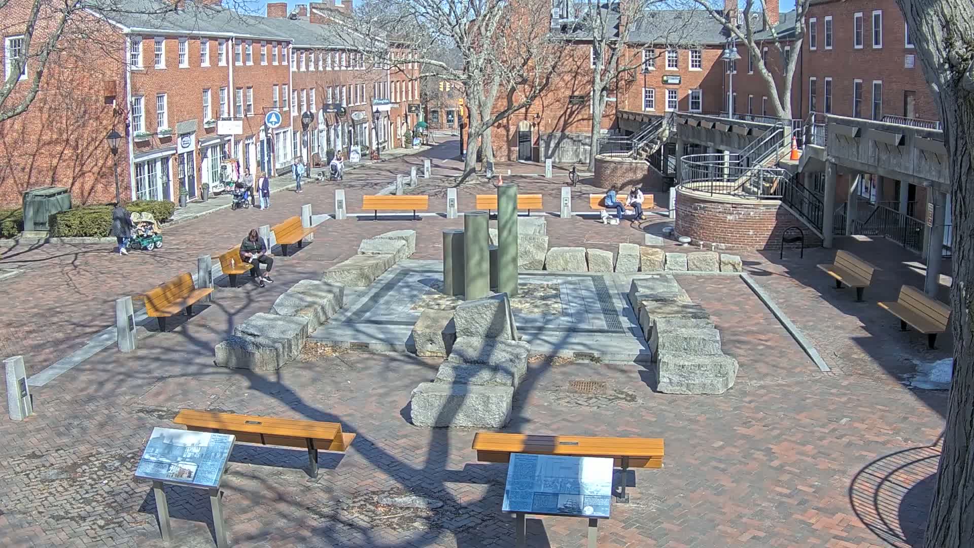 A brick plaza with several benches, a stone fountain, and brick buildings surrounds it on a sunny day.