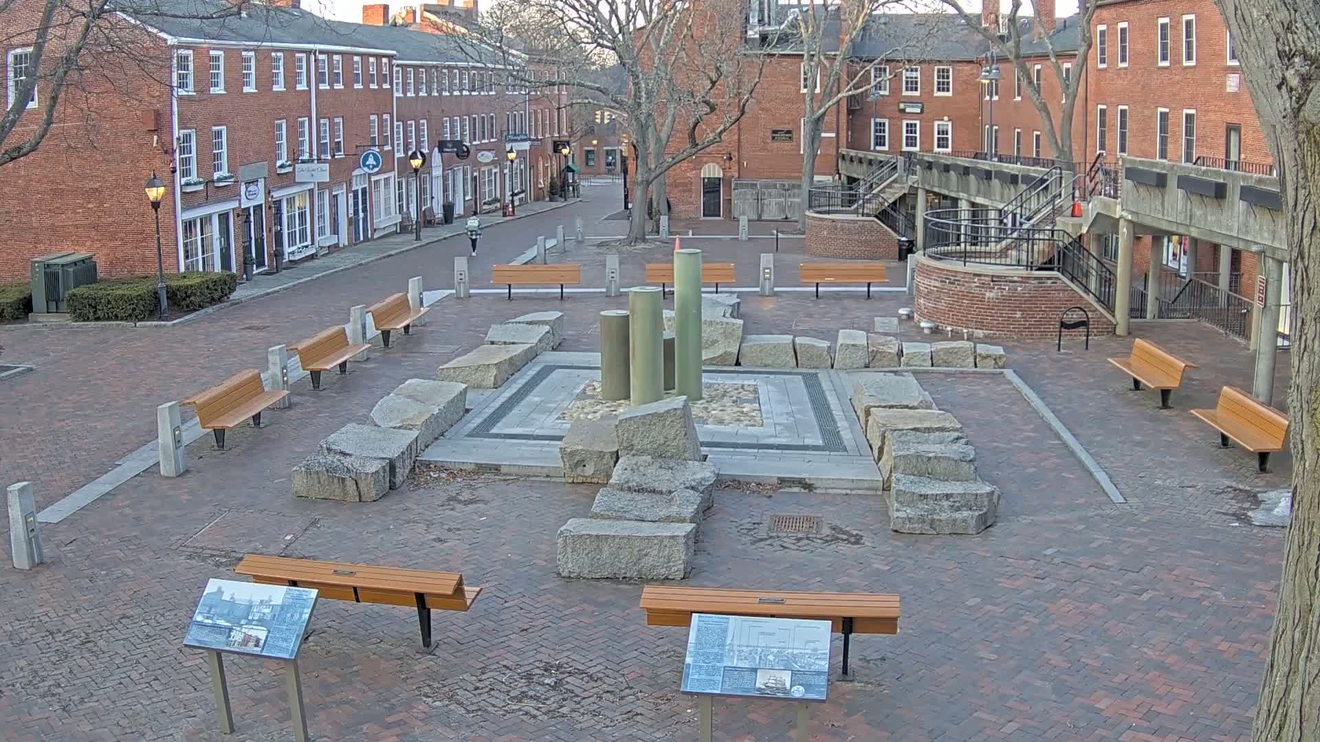 A brick plaza with several benches surrounds a stone and metal fountain, situated between brick buildings under a mostly clear sky.