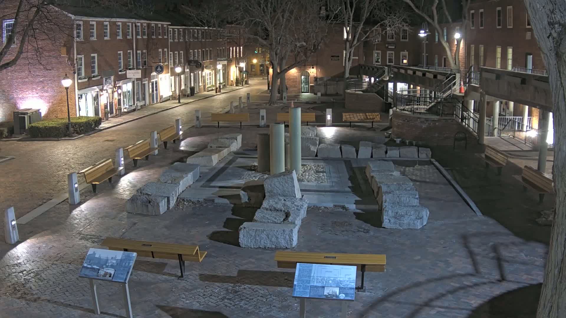 At night, a brick-paved plaza features a central area with large stone remnants and several modern benches, surrounded by brick buildings under the cover of trees.