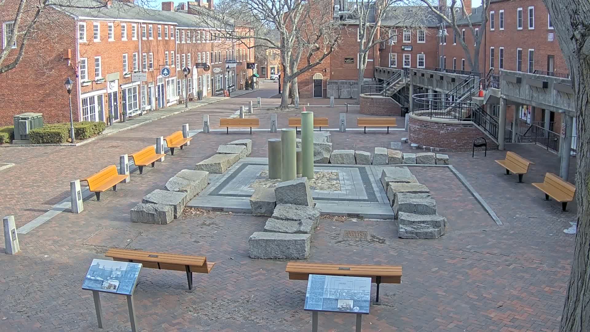 A brick plaza with several benches surrounds a stone fountain, situated between brick buildings on a sunny day.