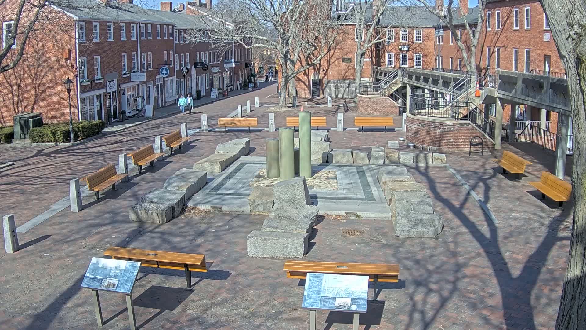 A brick plaza with several benches, a central stone structure featuring large, broken columns, and surrounding brick buildings under a sunny sky.