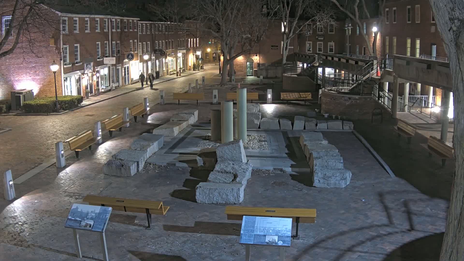 A nighttime view of a brick-paved plaza with remnants of stone columns and several benches, surrounded by brick buildings under a dark sky.