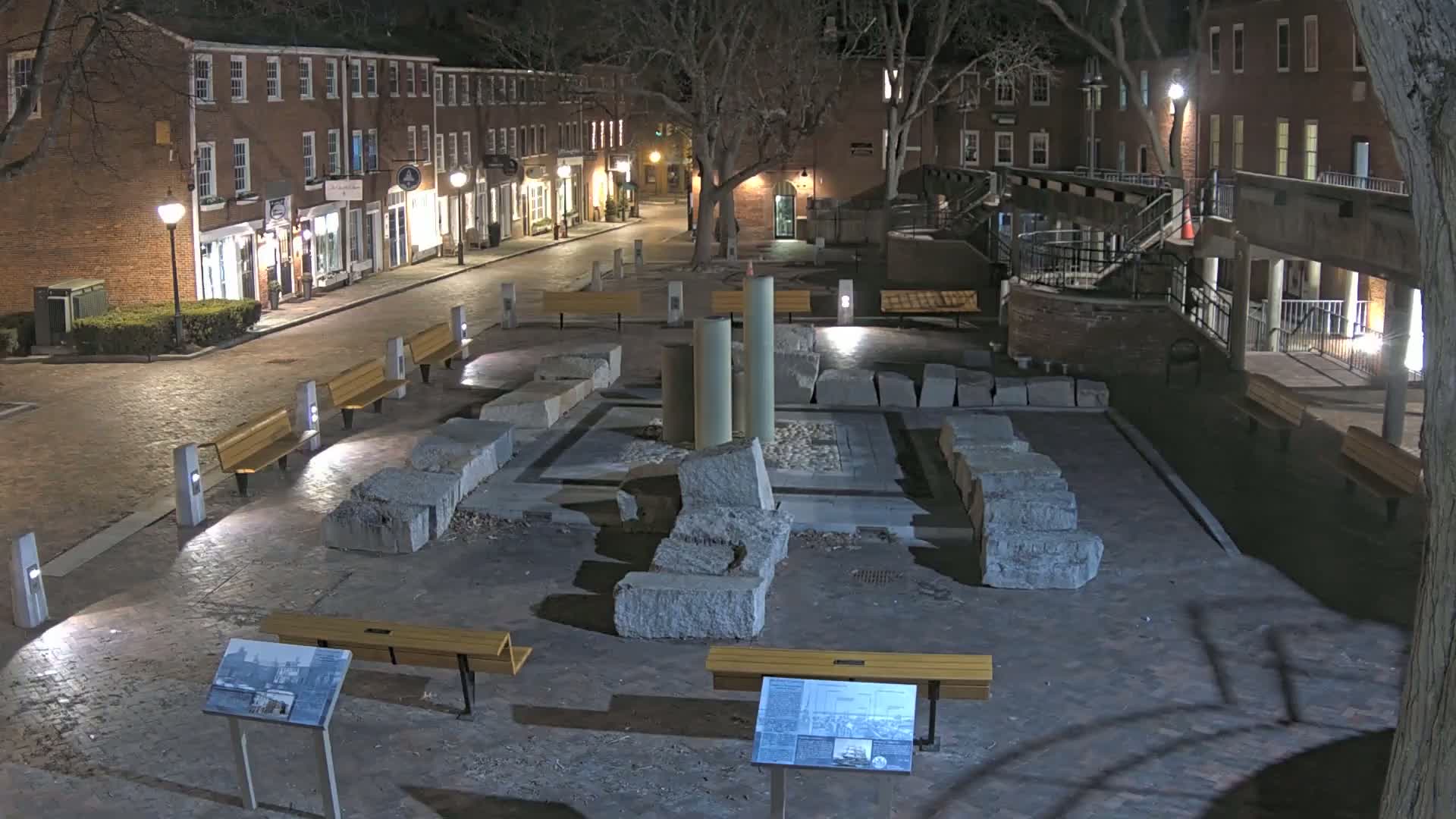 At night, a brick-paved plaza with several benches surrounds the ruins of a stone structure, illuminated by streetlights.