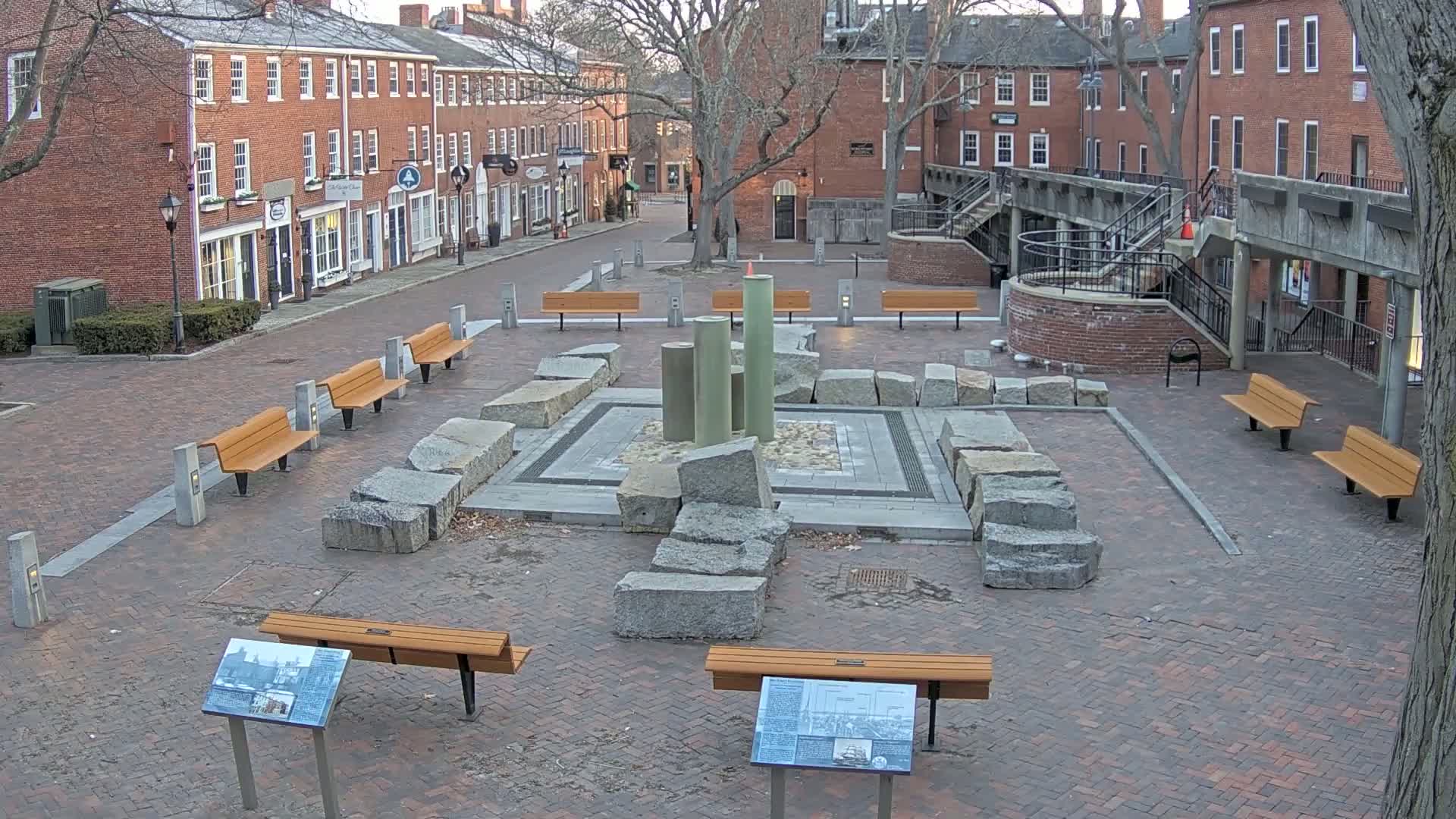 A brick plaza with several benches surrounds a stone fountain, situated between two rows of brick buildings under a mostly clear sky.