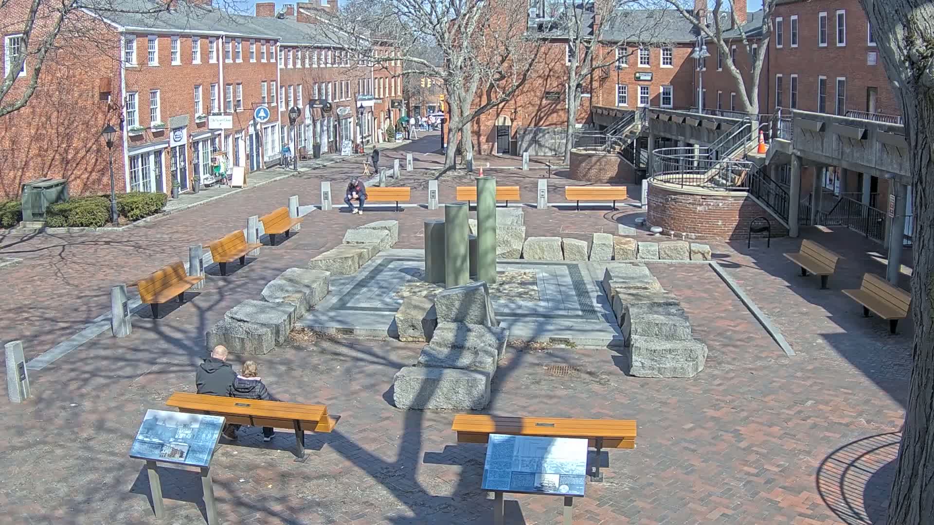 A brick plaza with a stone and metal fountain, several wooden benches, and informational signs sits between brick buildings on a sunny day.