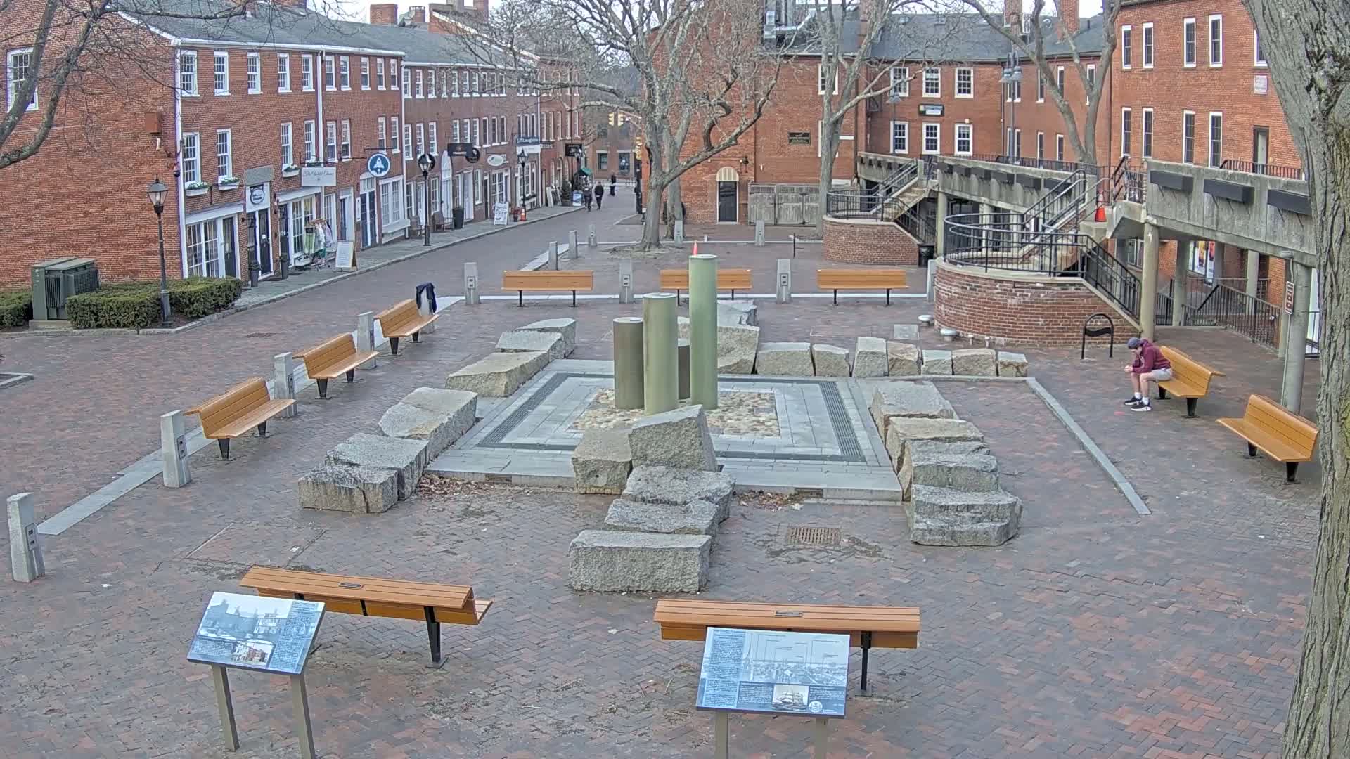 A brick plaza with several benches surrounds a stone and metal fountain, under a mostly clear sky, with brick buildings lining the street beyond.
