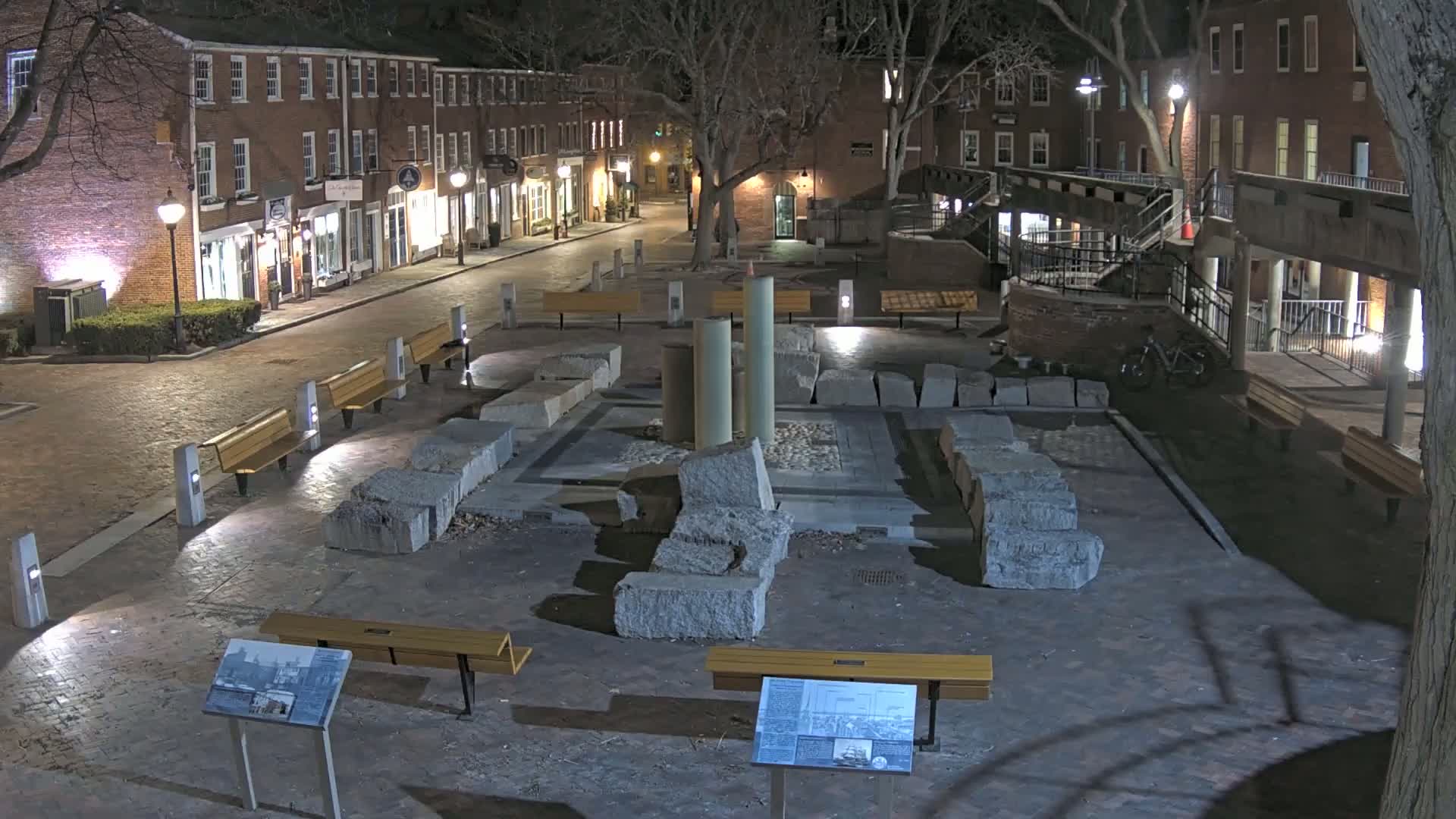 At night, a brick-lined street is illuminated, leading to a square containing the remnants of a stone structure, surrounded by benches and trees.