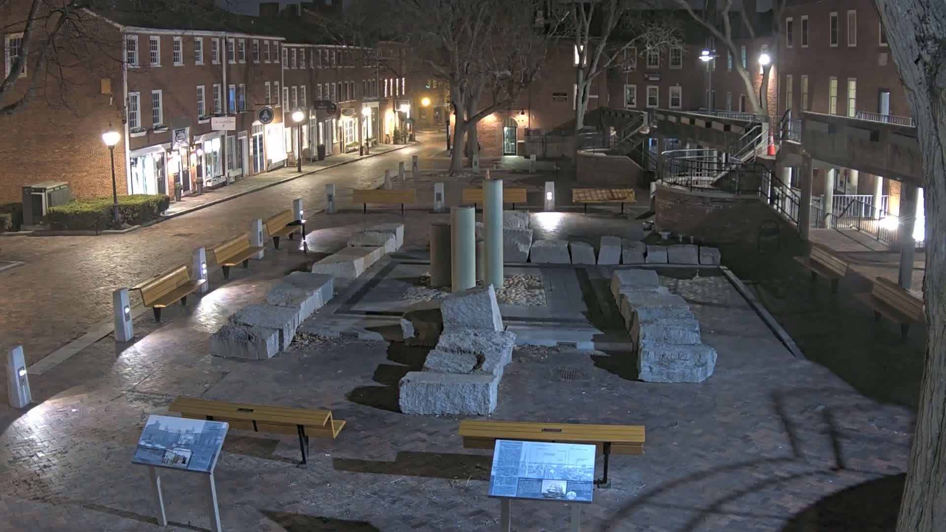 At night, a brick-lined town square features a central display of large stone blocks and several benches under lamplight.