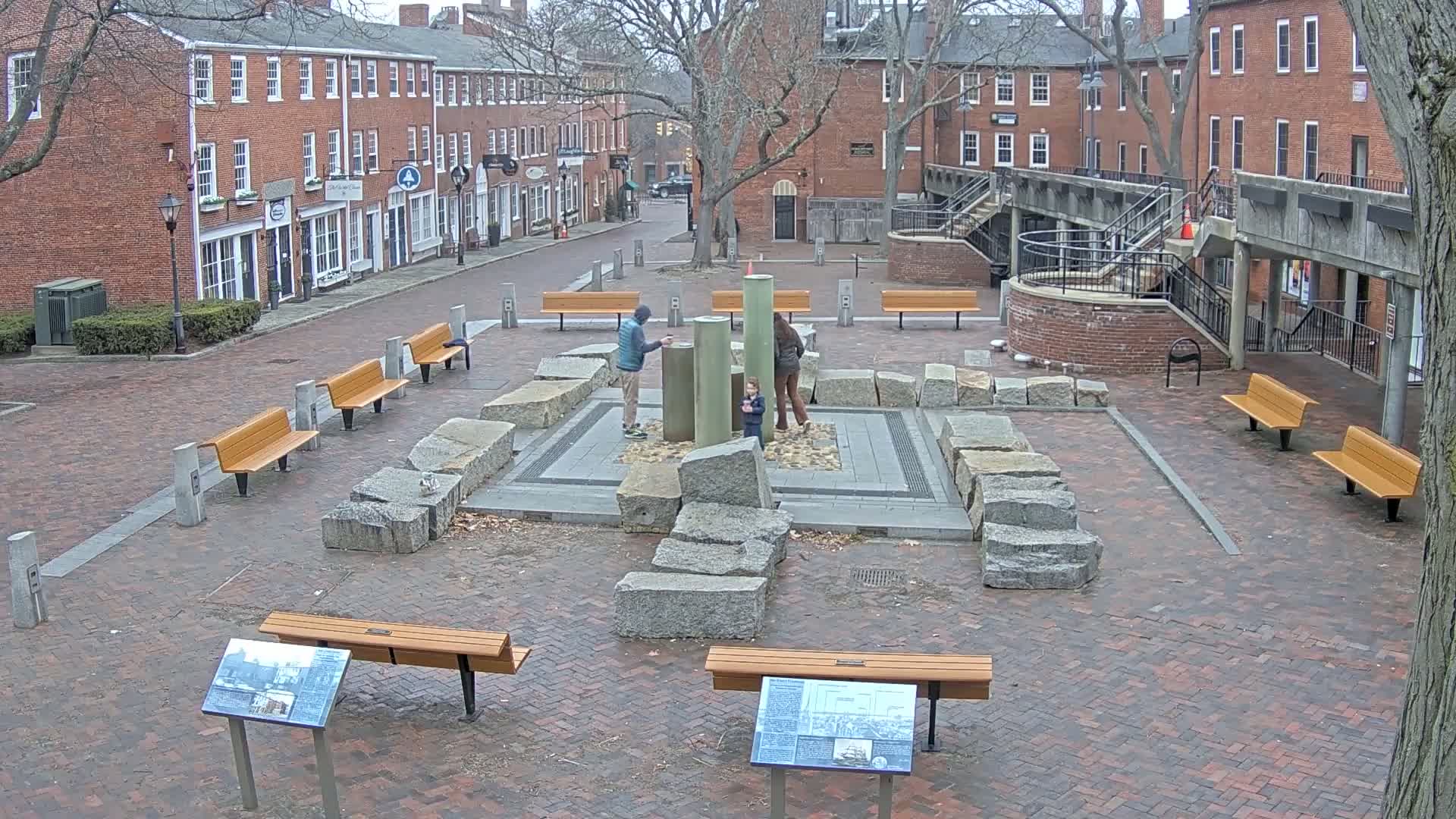 A brick plaza with a stone and metal water feature, surrounded by benches and brick buildings, on a cloudy day.