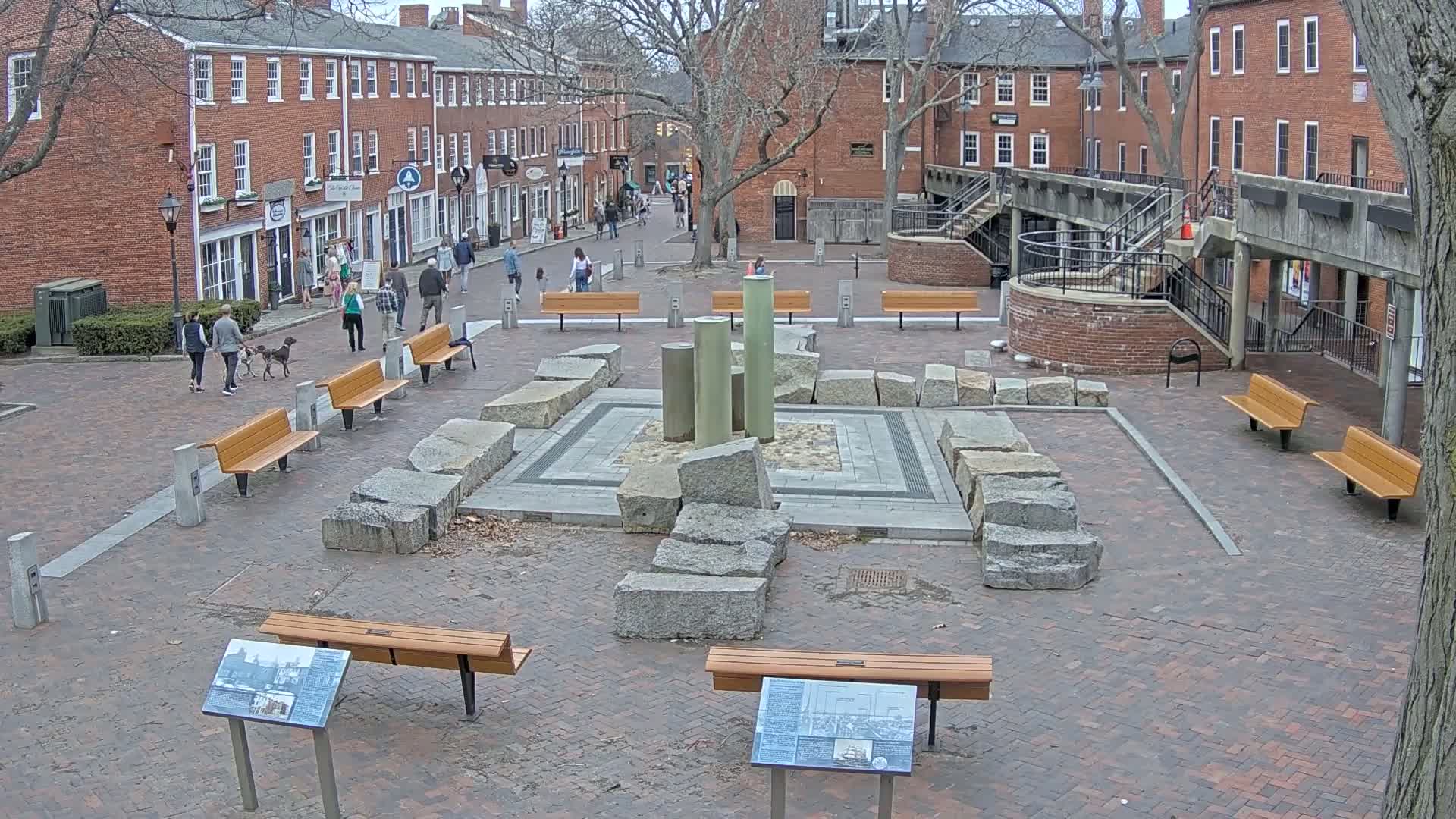 A brick plaza with benches and a central stone fountain, surrounded by brick buildings under a partly cloudy sky.