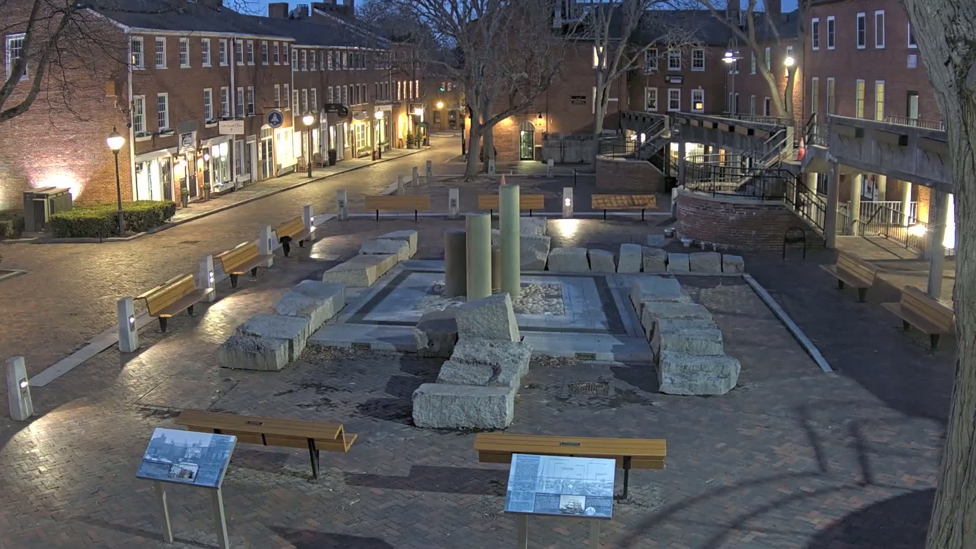 A brick plaza at night, featuring a stone structure in the center surrounded by benches and lit by streetlights, with brick buildings lining the street.