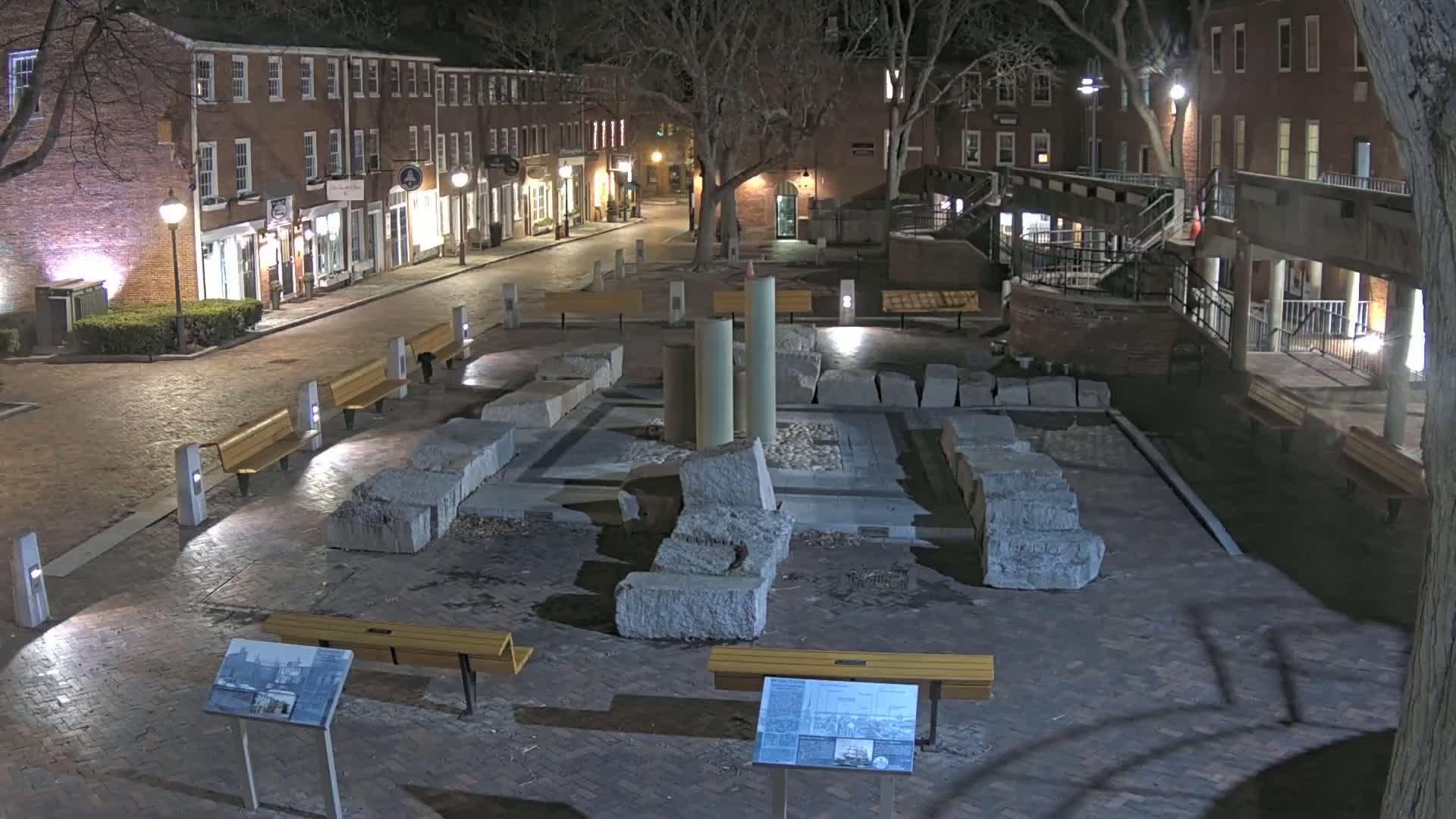 A nighttime view of a brick plaza with several benches, a stone structure resembling ancient ruins, and brick buildings lining a street in the background.