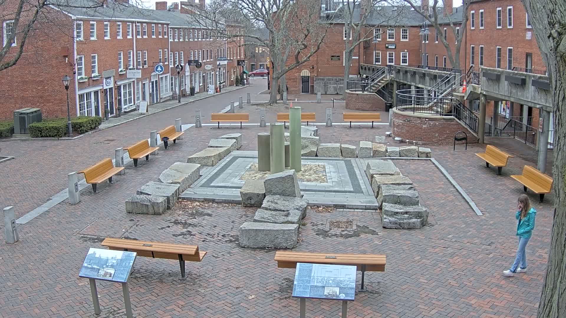 A brick plaza with several benches surrounds a stone and metal fountain, under a mostly cloudy sky.