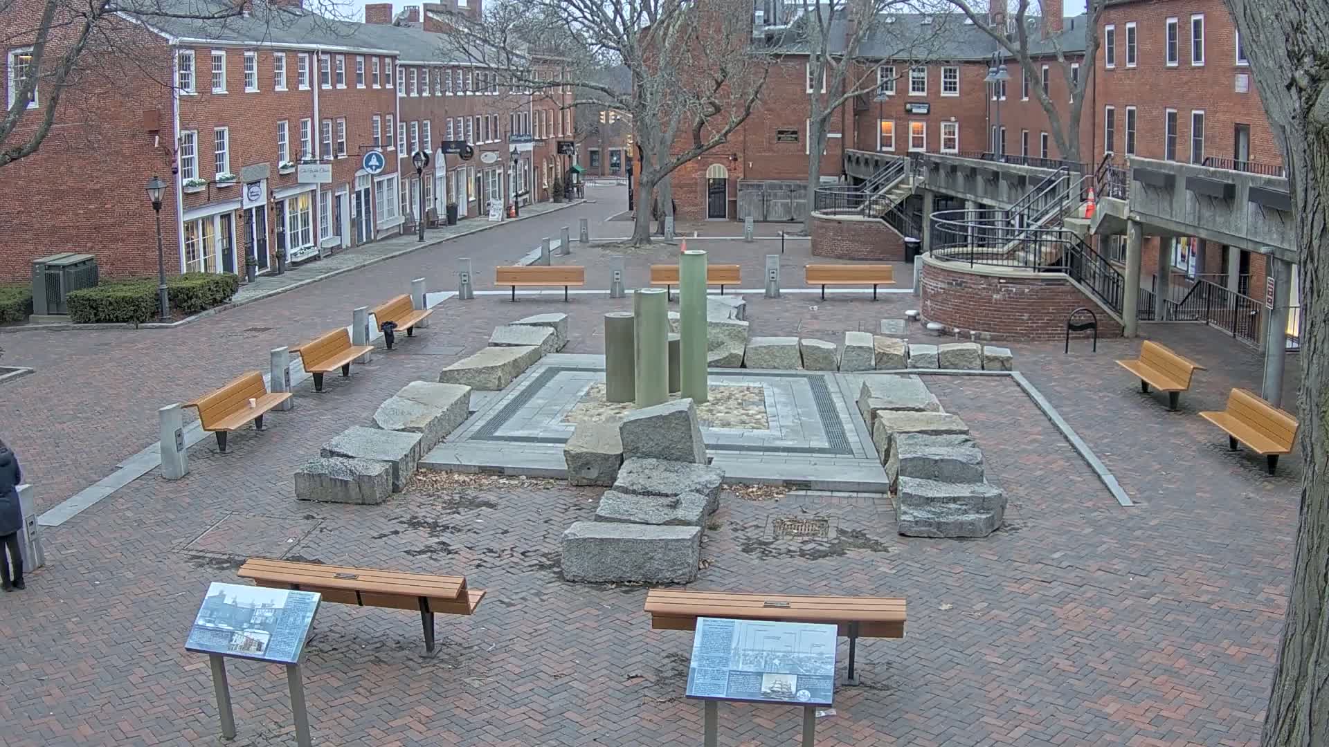 A brick plaza with benches surrounds a stone fountain, all situated between brick buildings on an overcast day.