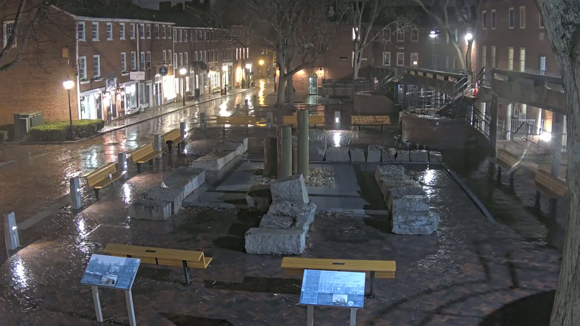 A rain-slicked brick plaza at night features a stone monument surrounded by benches and lit by streetlights, with brick buildings visible in the background.