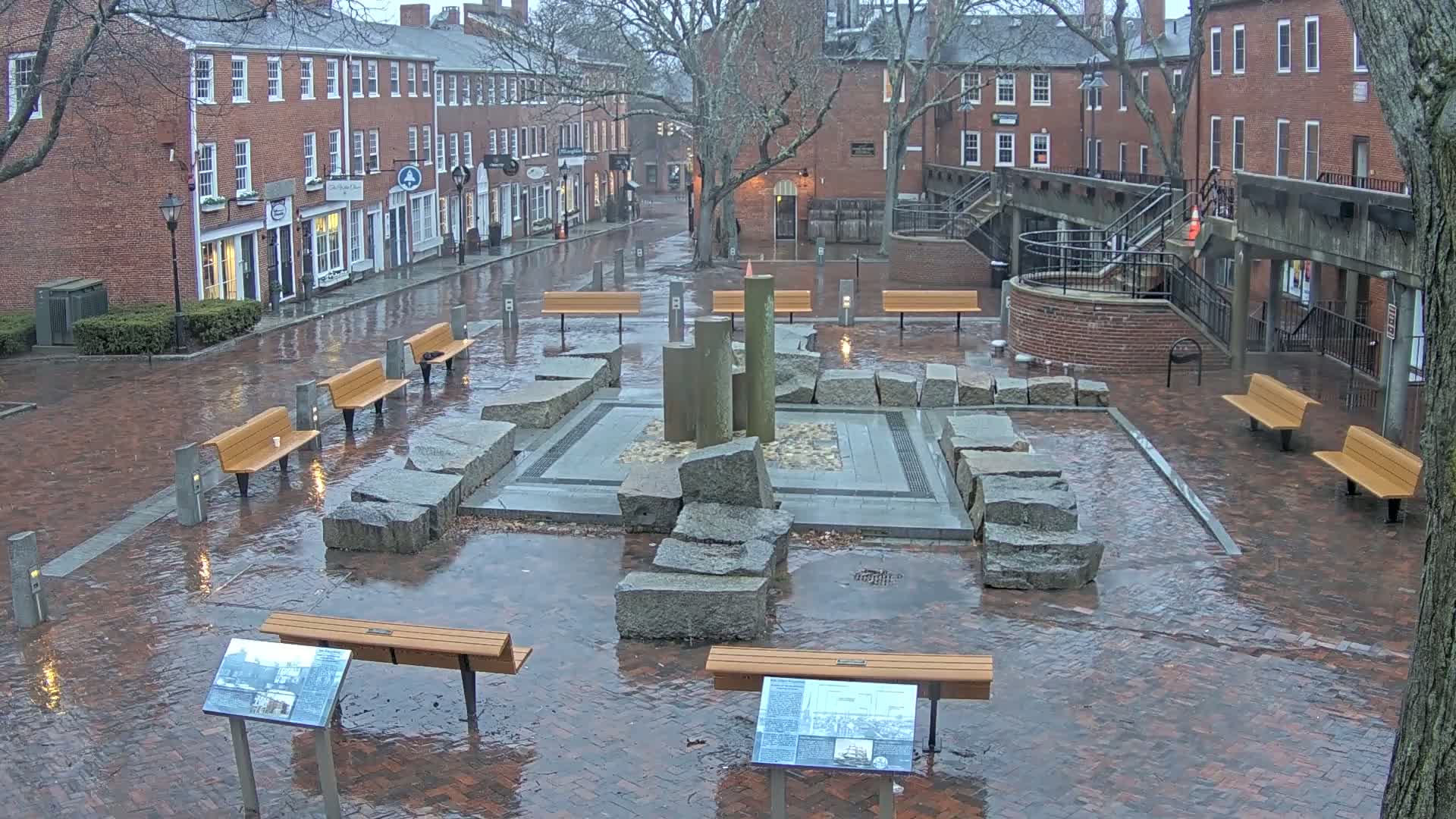 A rain-soaked brick plaza features a stone fountain, several benches, and historical markers, situated between rows of brick buildings under a gray sky.