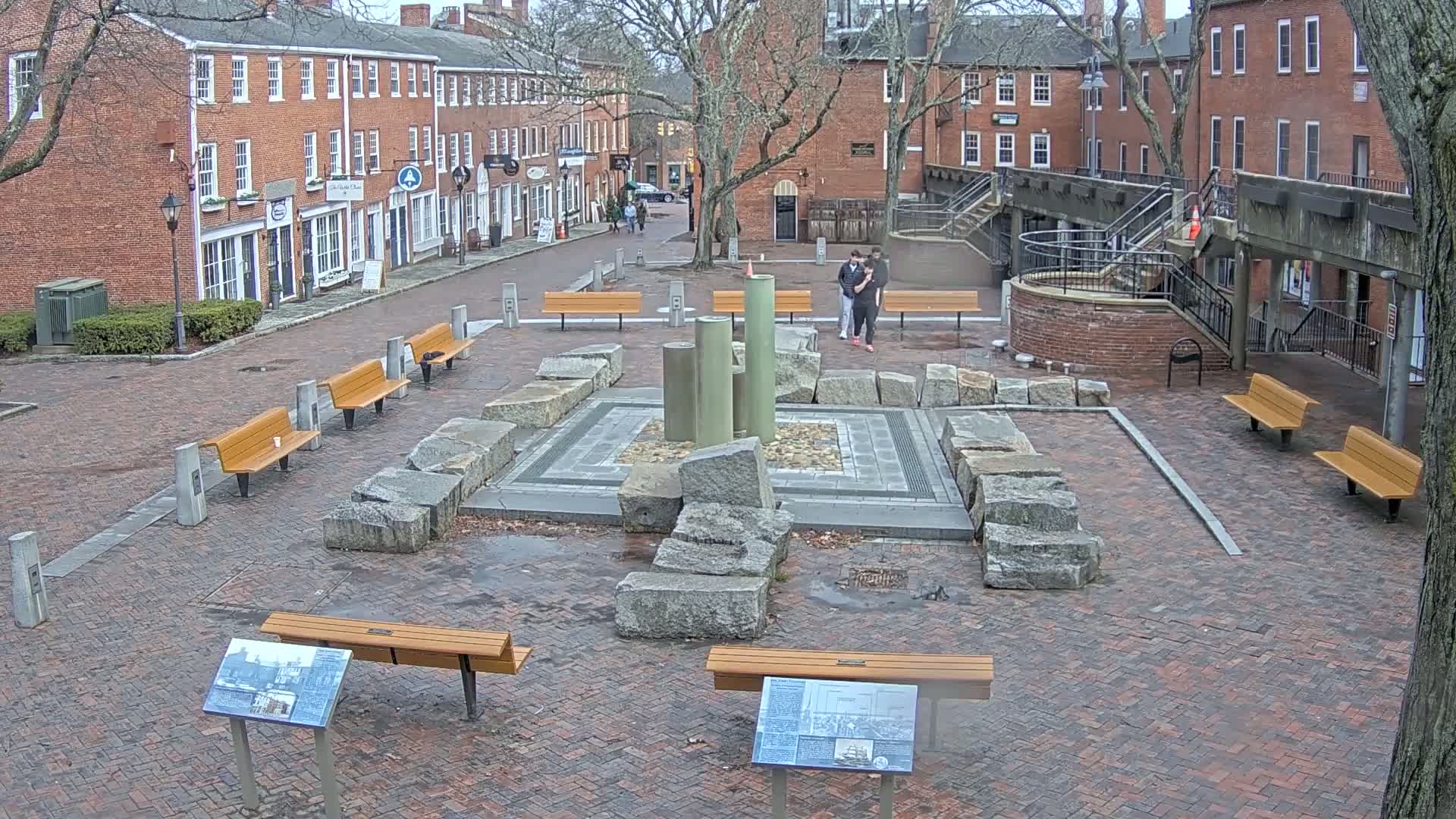 A brick plaza with several benches surrounds a stone fountain, and a couple stands nearby in front of brick buildings under an overcast sky.