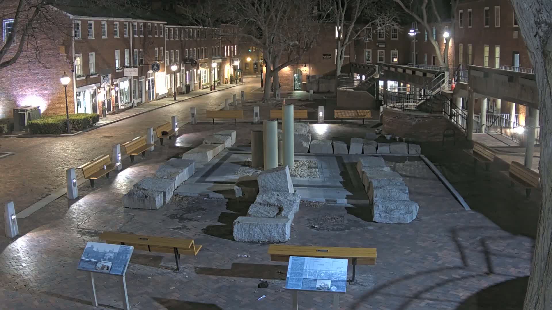 At night, a brick plaza contains a stone structure with several large blocks and columns, surrounded by benches and streetlights, with brick buildings lining the street behind it.