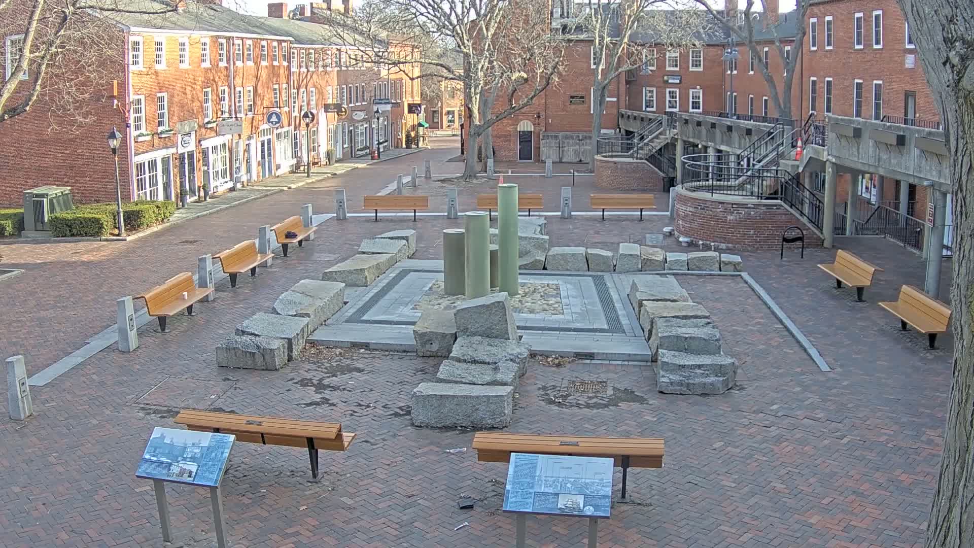 A brick plaza with several benches surrounding a stone and metal fountain-like structure, situated between brick buildings under a mostly clear sky.