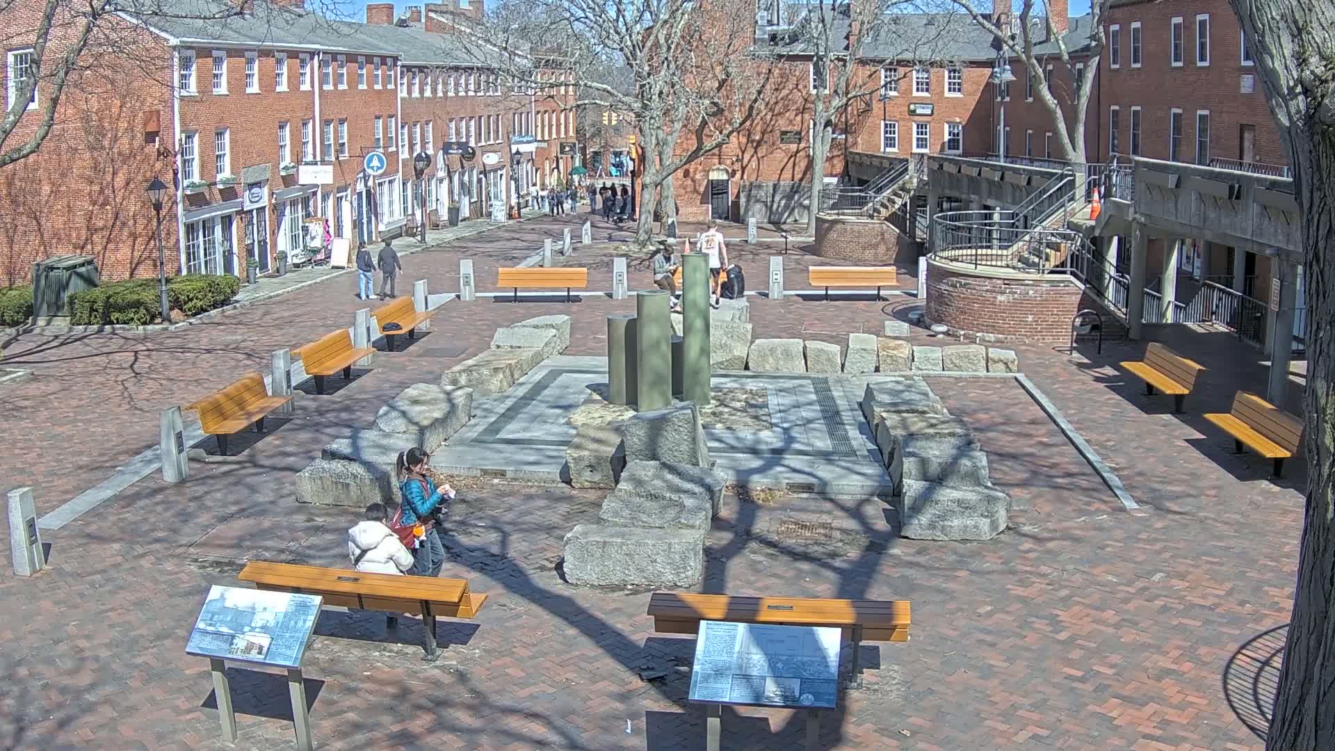 A brick plaza with several wooden benches surrounds a central stone structure, under a sunny sky with bare trees.