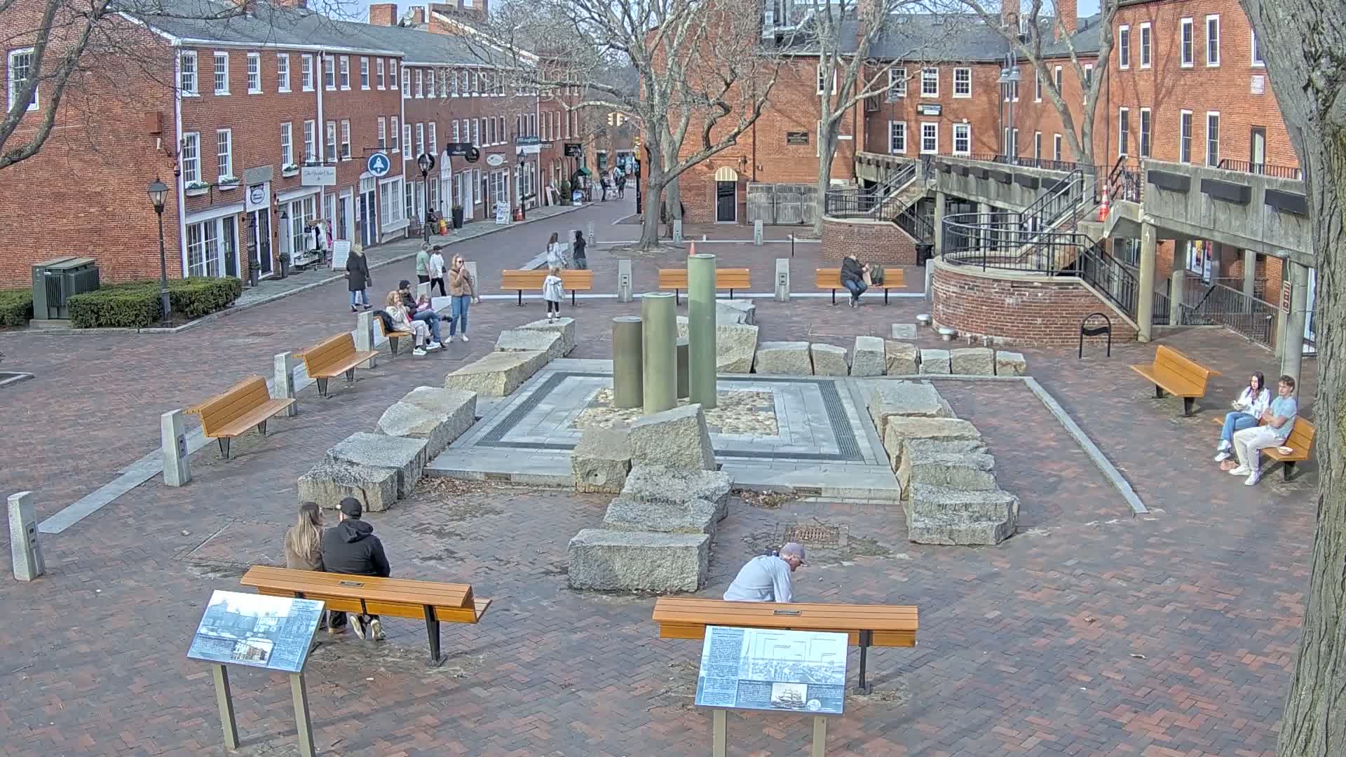 A brick plaza, surrounded by brick buildings, features a central stone structure with several people sitting on benches on a partly sunny day.