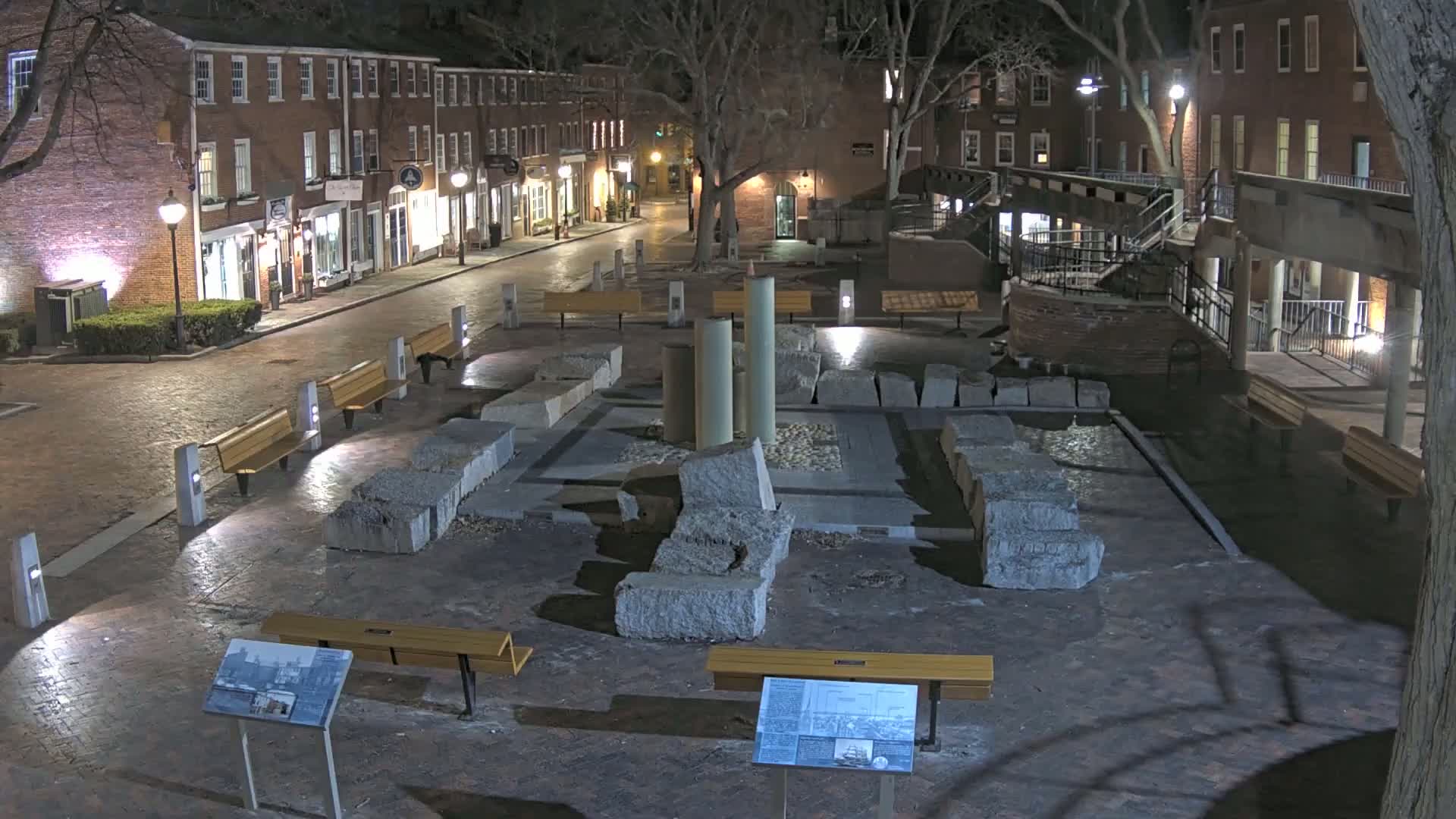 At night, a brick-lined plaza with several benches surrounds a central area featuring remnants of stone columns and walls, illuminated by streetlights.
