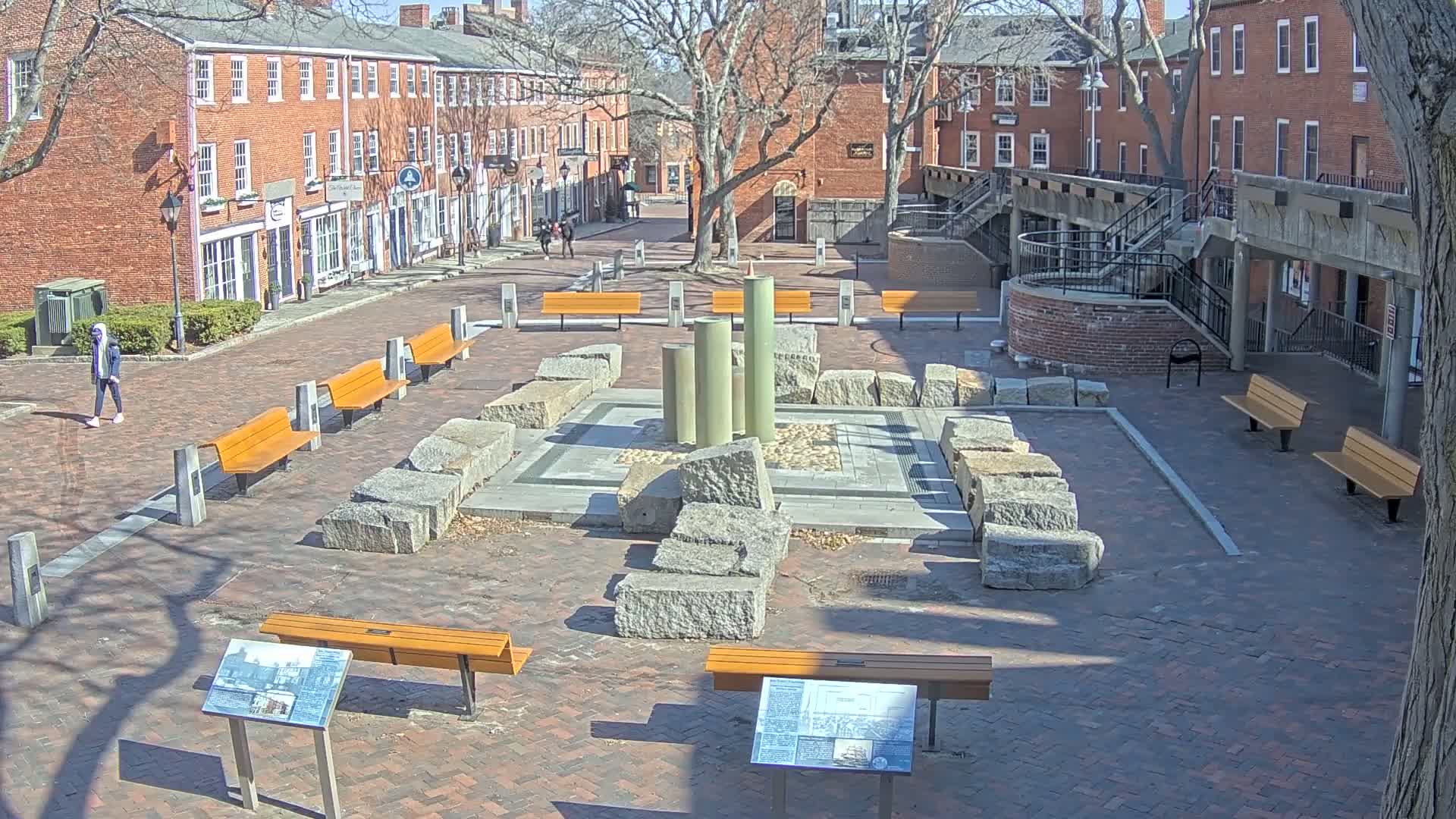 A brick plaza with several benches, a stone fountain, and surrounding brick buildings under a sunny sky.