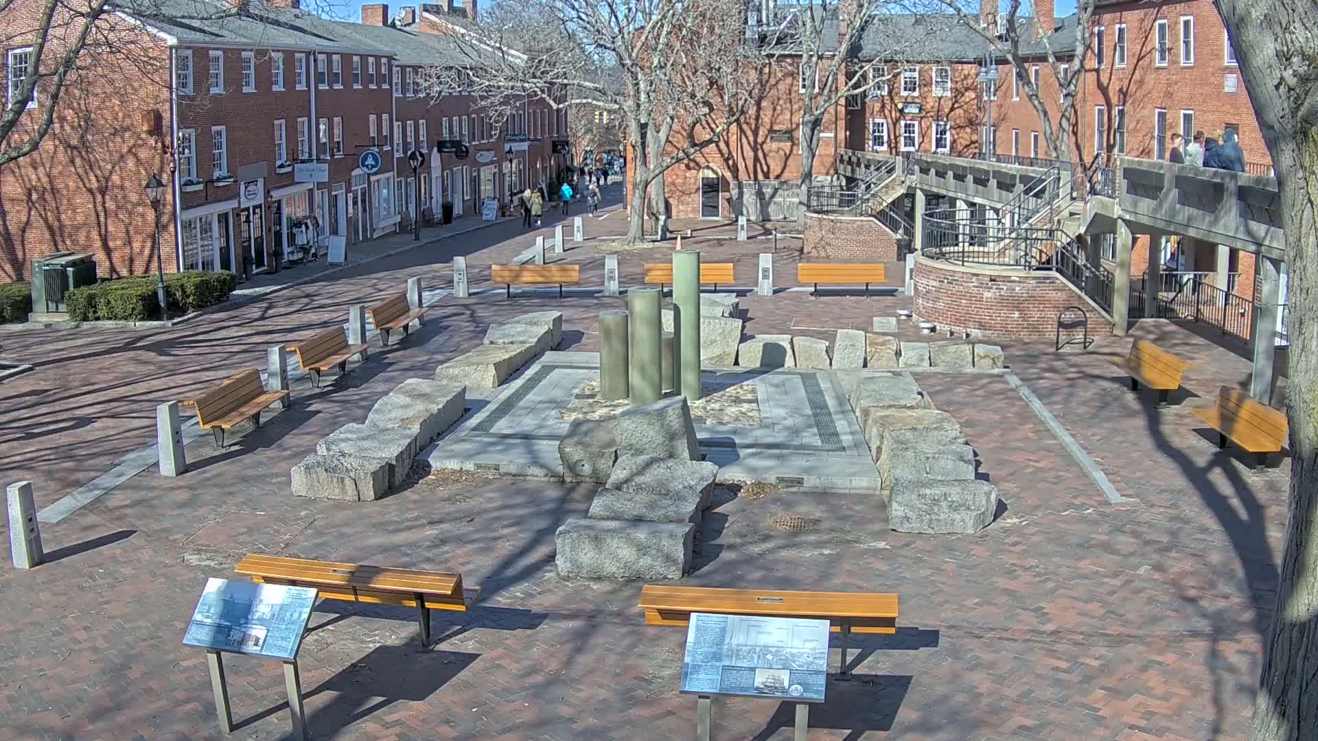 A brick plaza with several benches, a central stone structure, and surrounding brick buildings under a sunny sky.