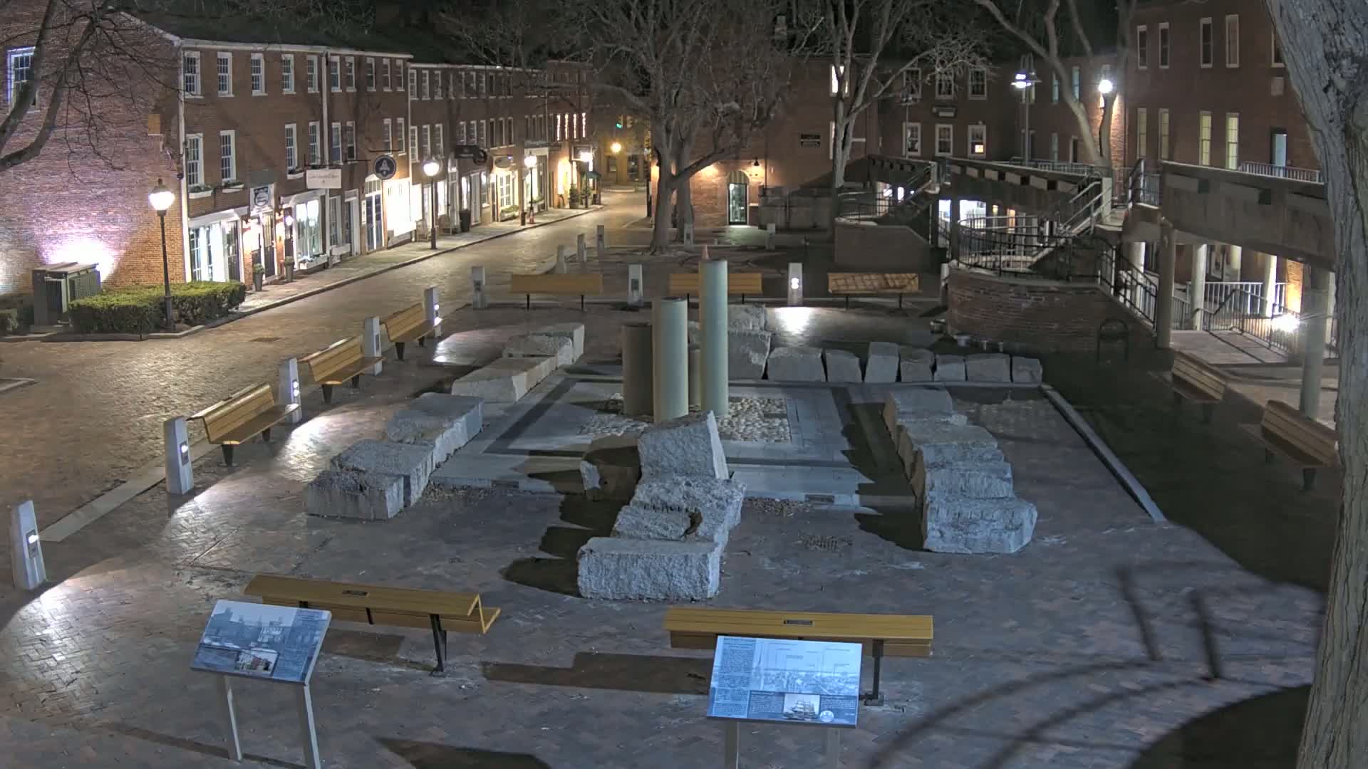 At night, a brick-paved plaza features a stone ruin in the center surrounded by benches and lit by streetlights, with brick buildings lining a street visible in the background.