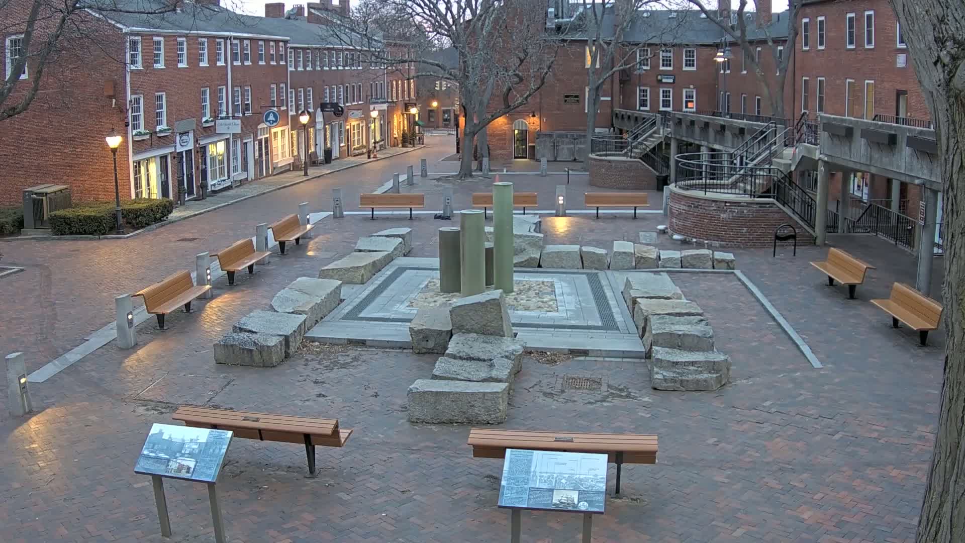 A brick plaza at dusk features a central stone structure with several cylindrical columns, surrounded by numerous benches and informational plaques.