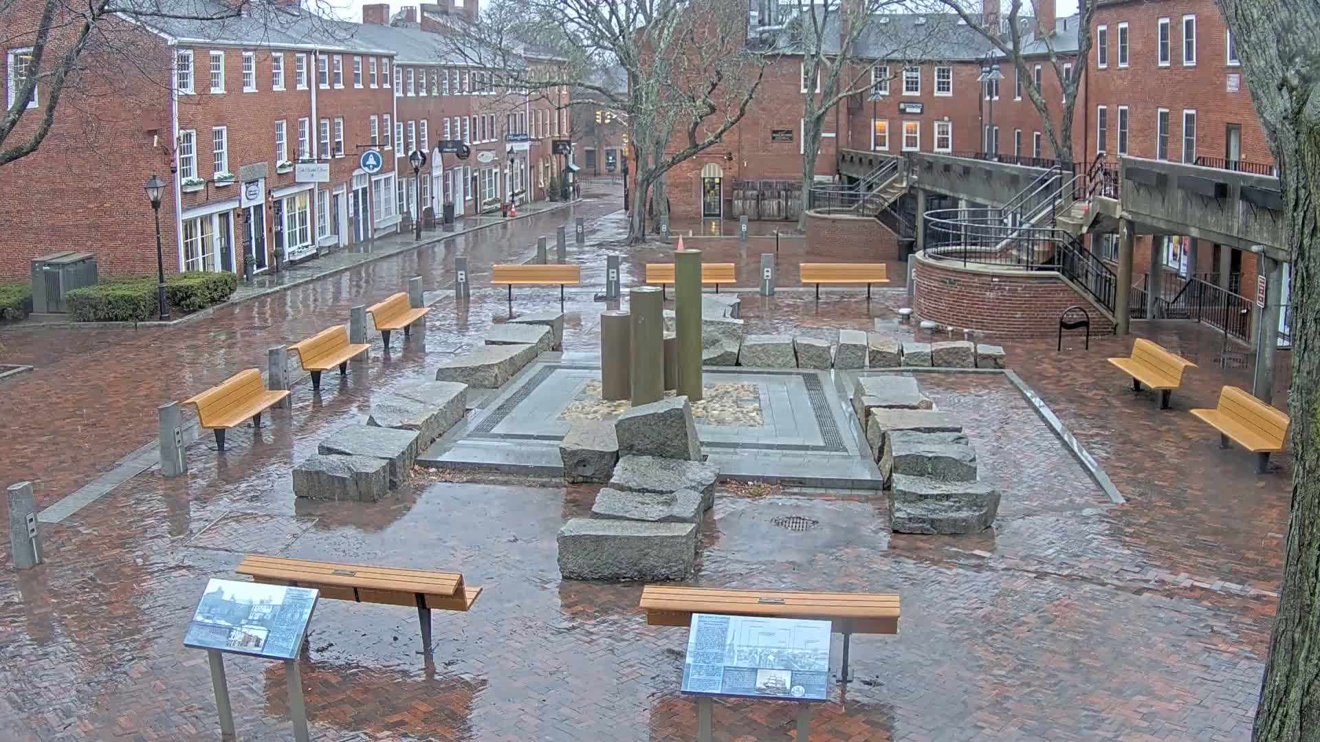 A wet brick plaza with several benches surrounds a stone fountain, located between red brick buildings under an overcast sky.