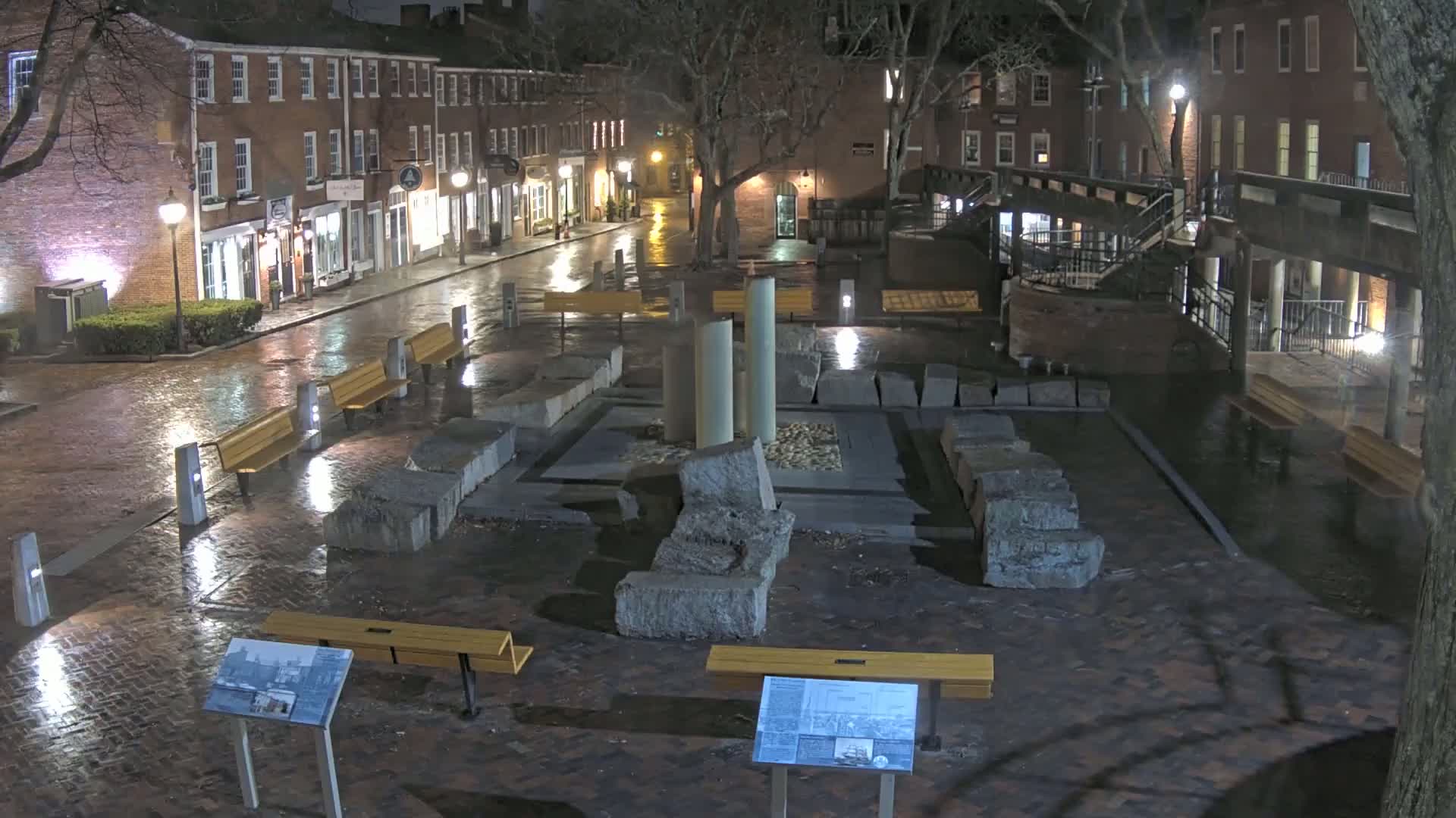 A wet, brick plaza at night features several benches, a stone structure, and a row of illuminated buildings in the background.
