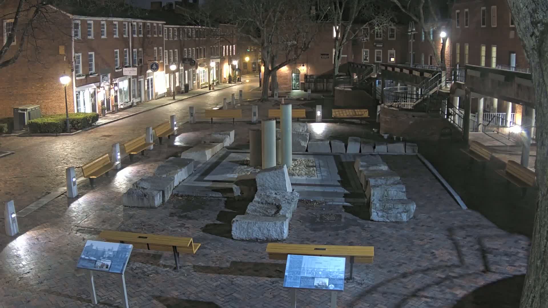 A nighttime view of a brick-paved plaza with stone ruins at its center, surrounded by brick buildings and illuminated by streetlights.