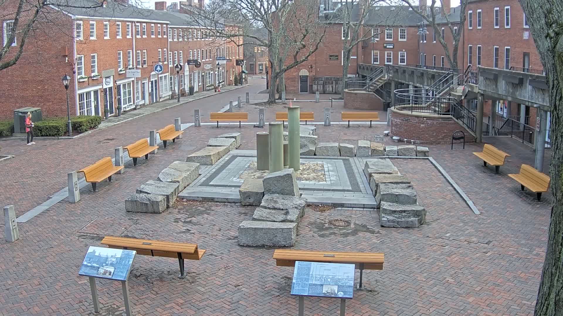 A brick plaza with several benches surrounds a stone fountain, overlooked by brick buildings under a cloudy sky.