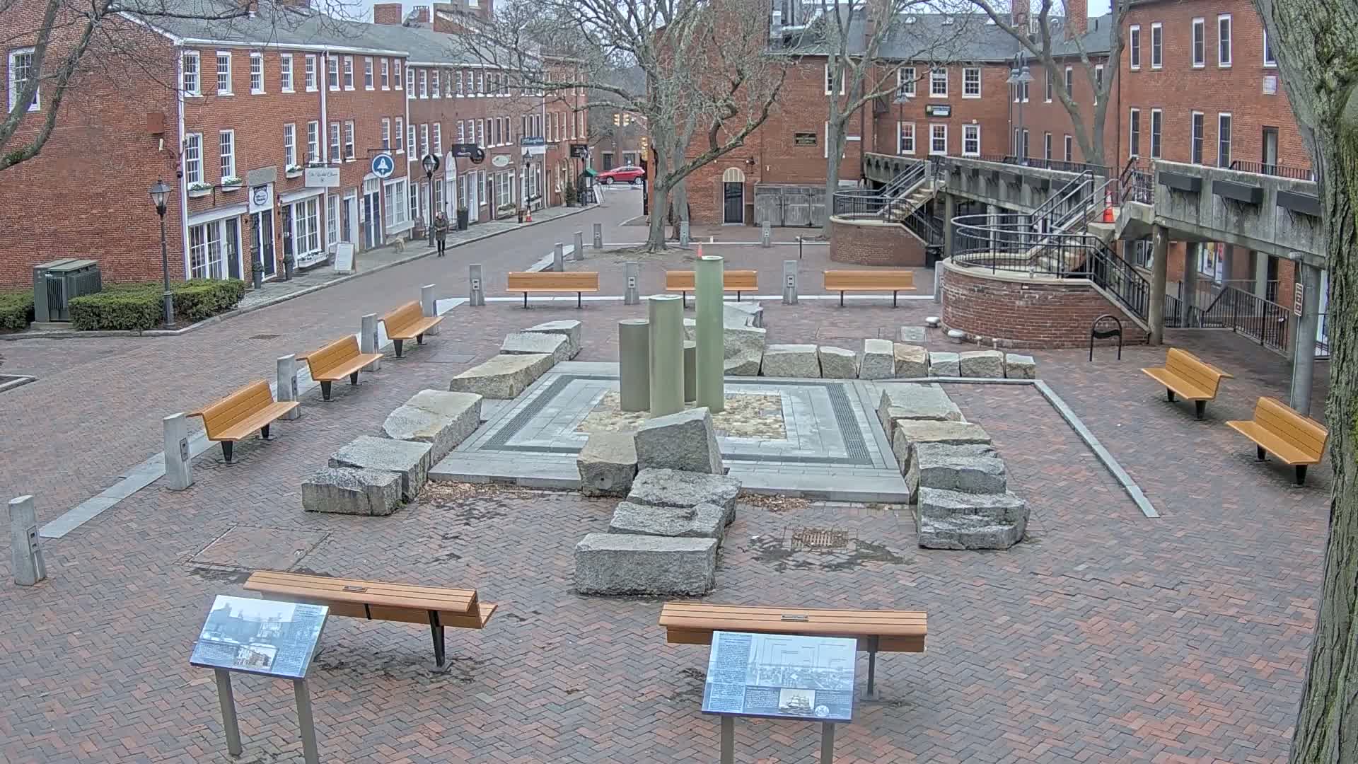 A brick plaza with several benches surrounds a stone and metal fountain-like structure, situated between brick buildings on an overcast day.