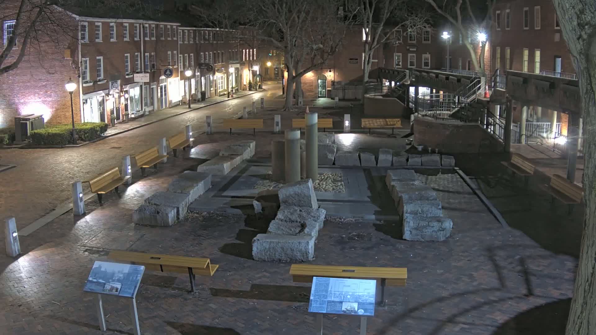 A nighttime scene of a brick plaza with stone remnants in the center, surrounded by benches and buildings with streetlights illuminating the area.